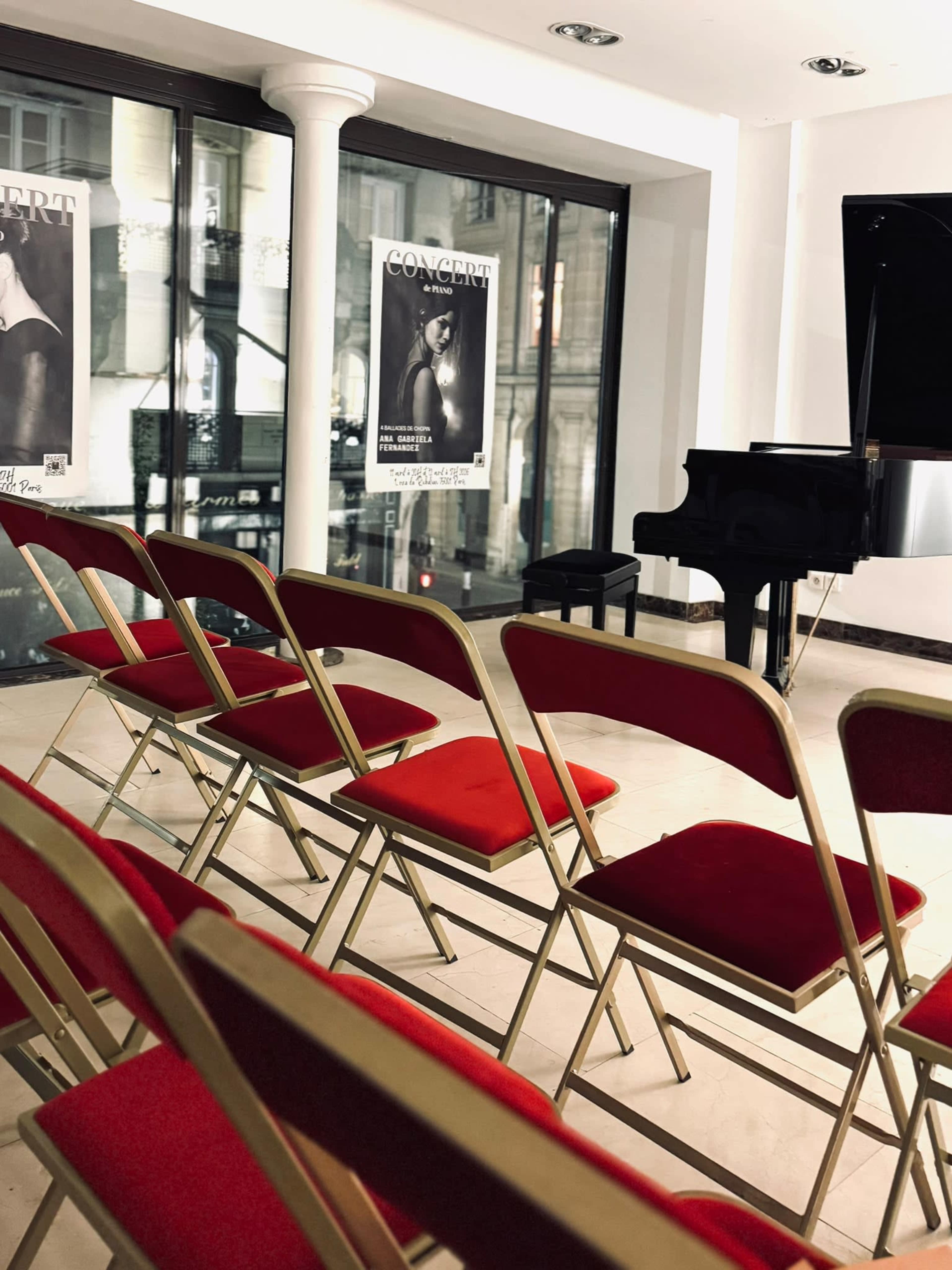 The image shows a row of red chairs facing a black piano in a small concert venue with posters on the walls.