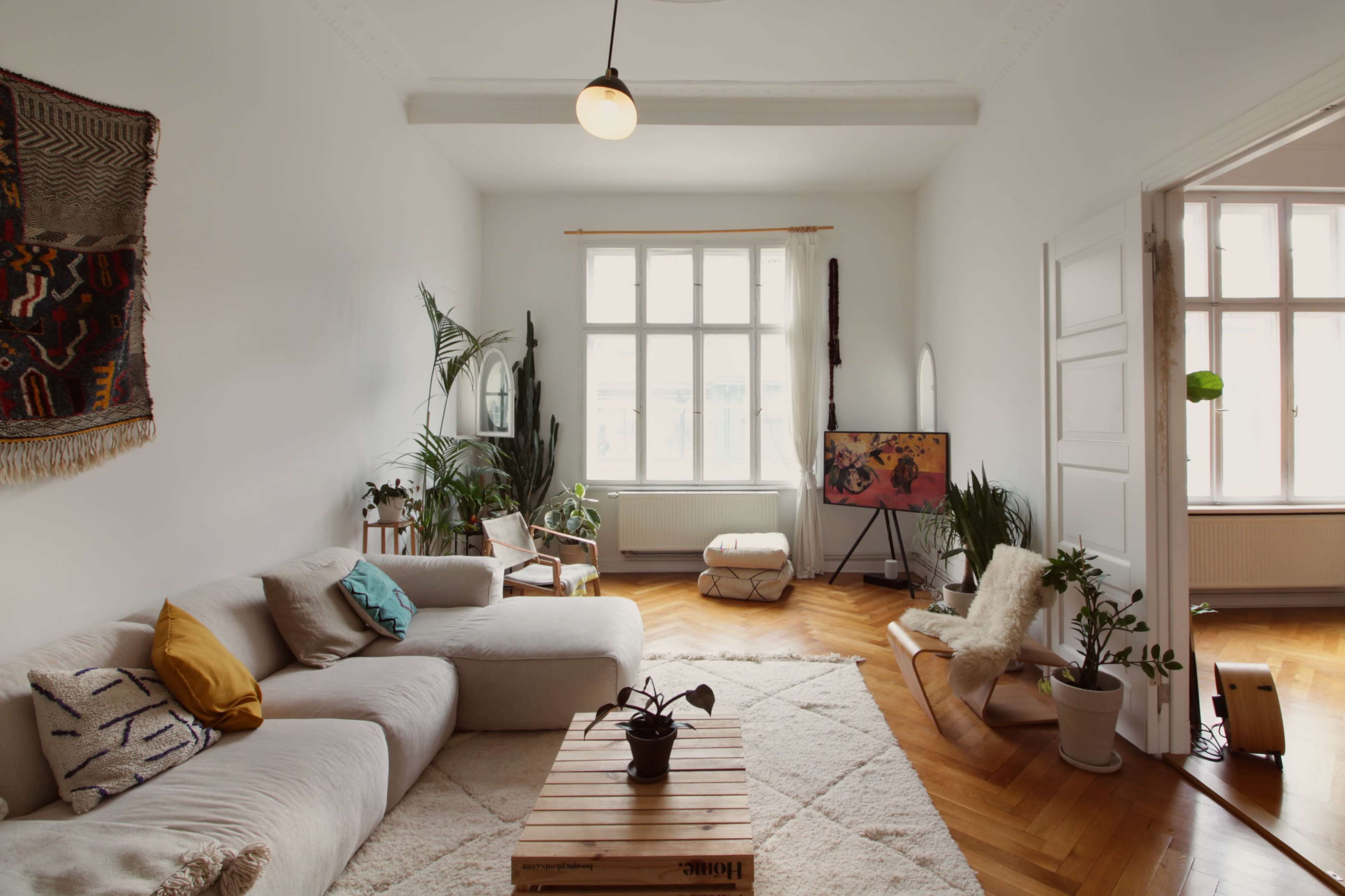 The image shows a brightly lit, minimalist living room featuring a light-colored sofa, wooden flooring, indoor plants, and a modern art piece on display.