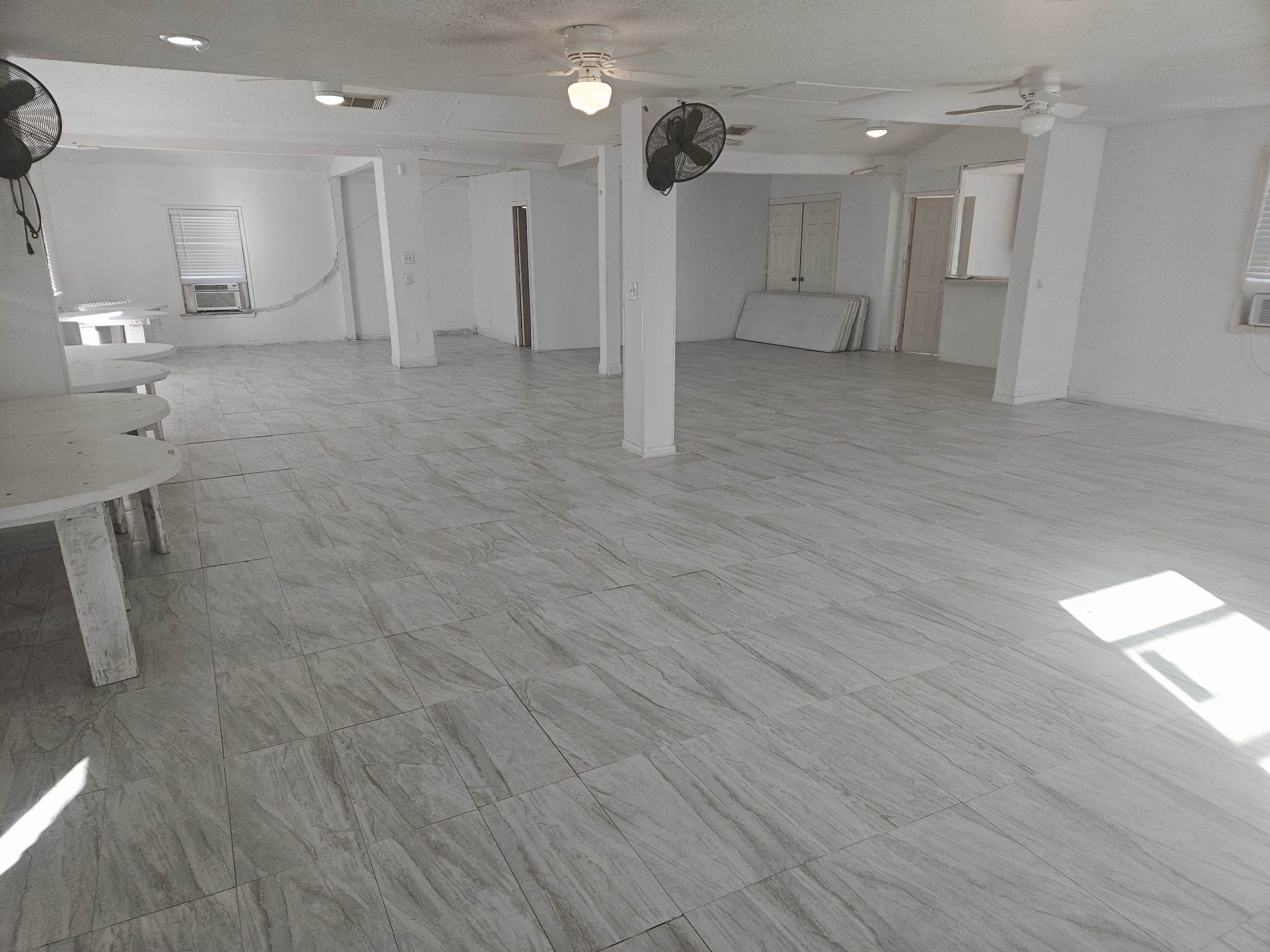 The image shows a spacious, empty room with tiled flooring, white walls, and several ceiling fans.