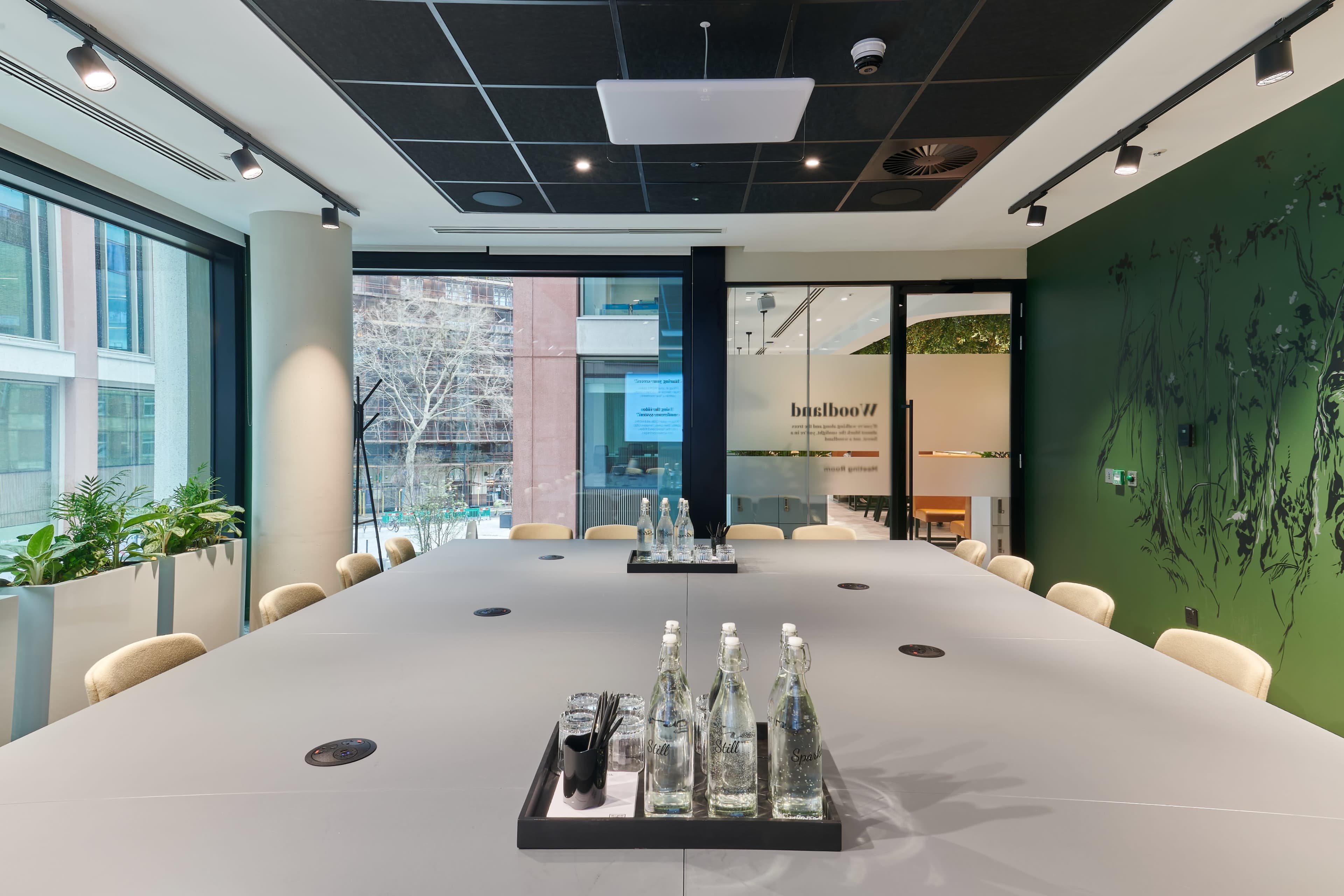 A modern conference room features a large table centered with water bottles and glasses, surrounded by windows that provide views of greenery outside.