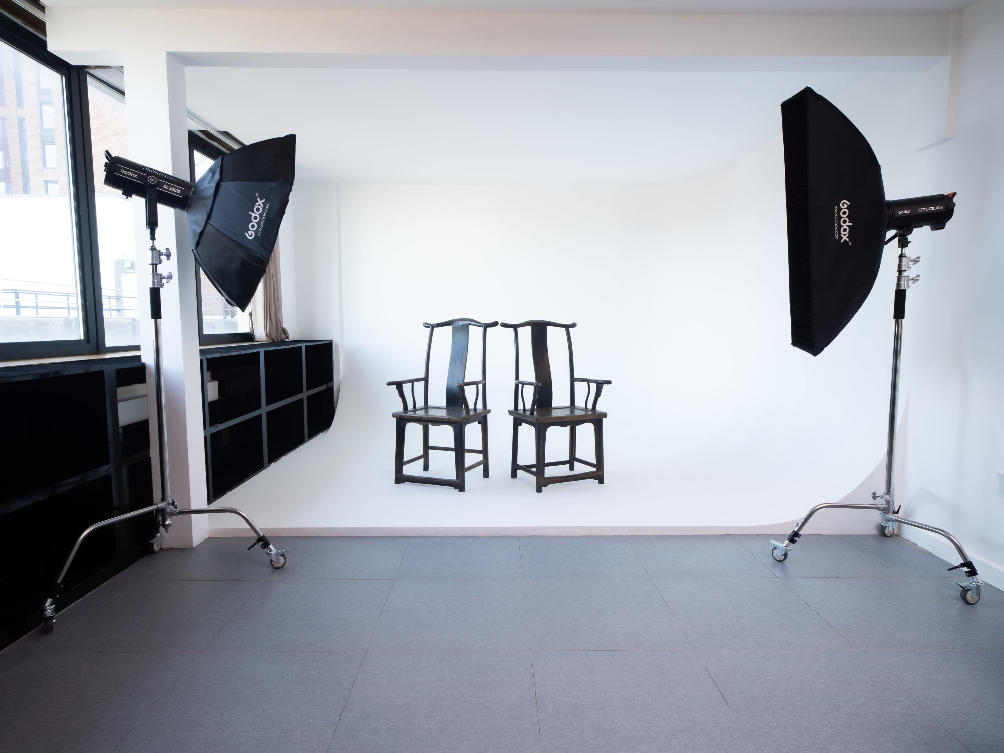 The image shows a minimalist studio setup with two wooden chairs placed in front of a blank white backdrop, flanked by studio lights on stands.