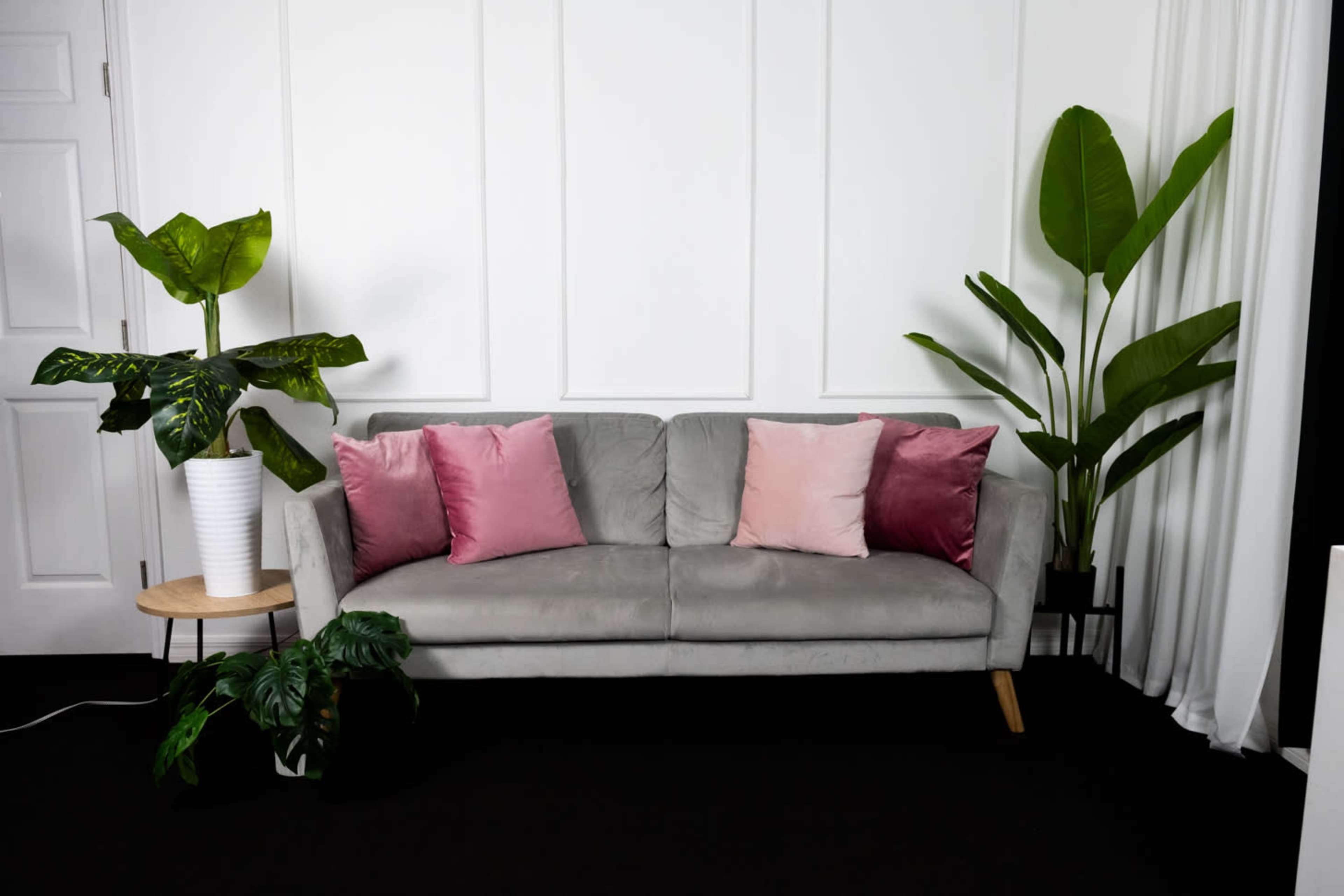 A gray sofa with pink and burgundy pillows is positioned against a white wall, flanked by potted plants on either side.