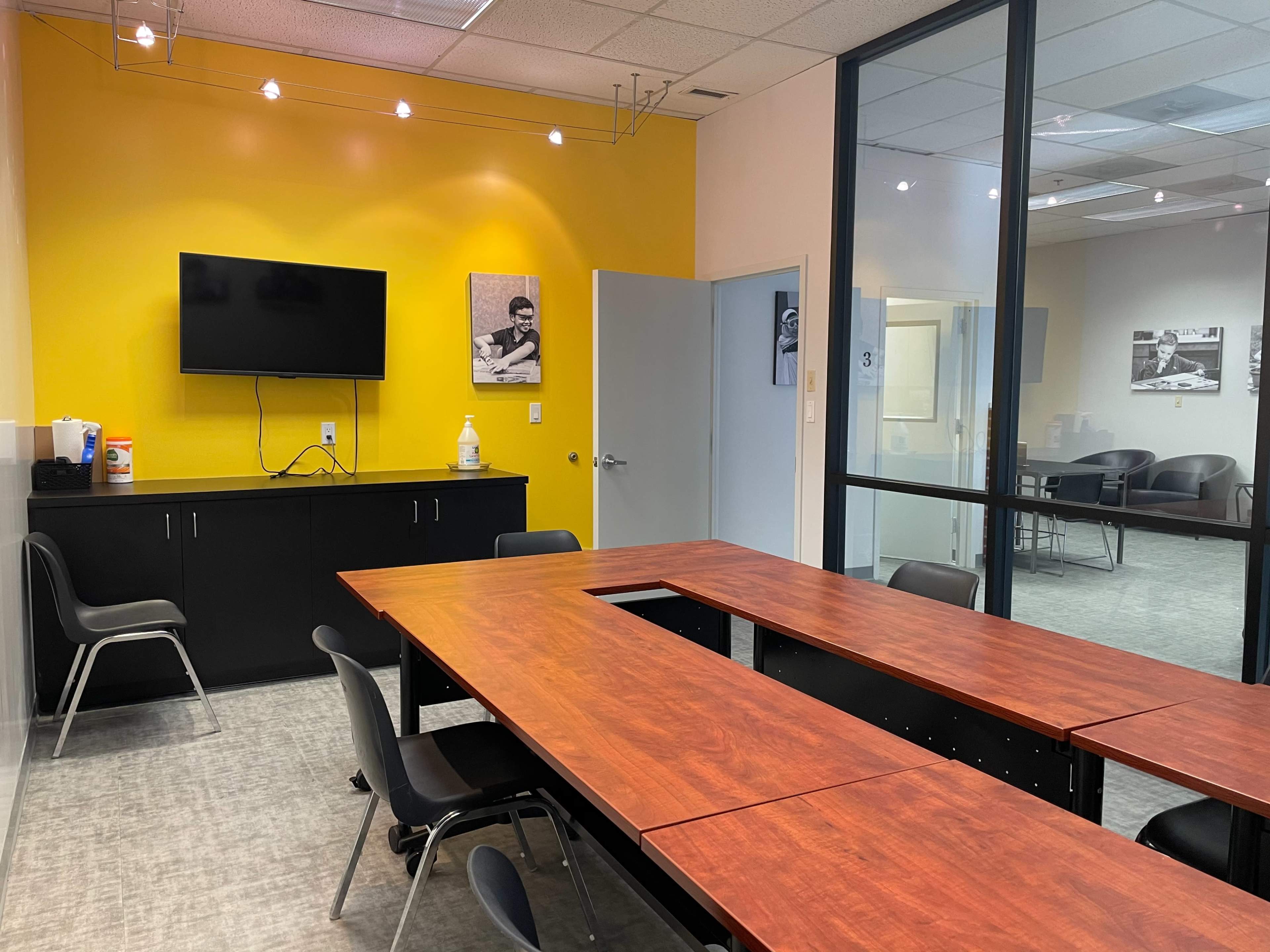 A conference room with a wooden table, black chairs, a television mounted on a yellow wall, and glass walls displaying another room in the background.