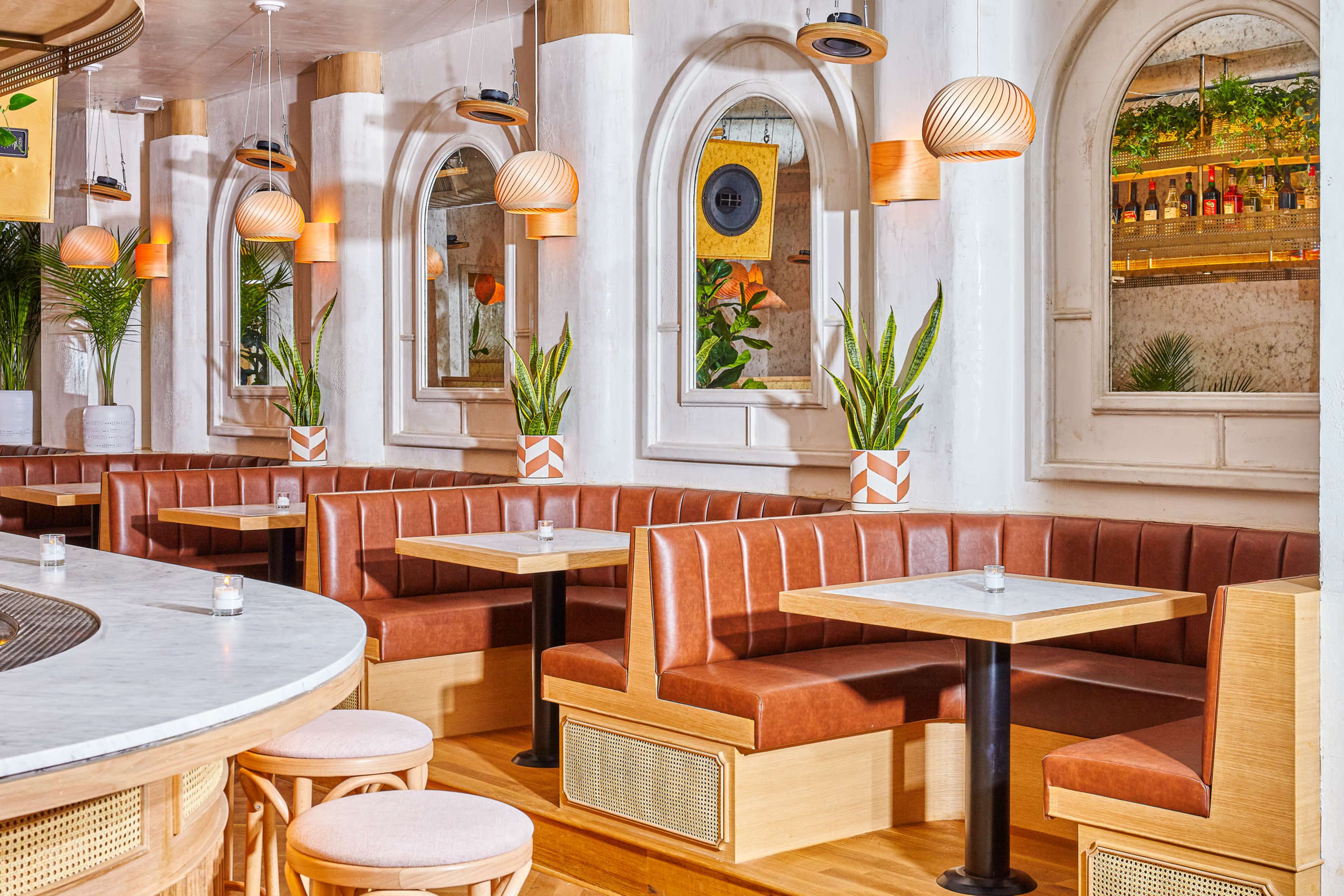 A modern dining space with leather banquette seating, white walls, and plants, illuminated by pendant lights above small tables.