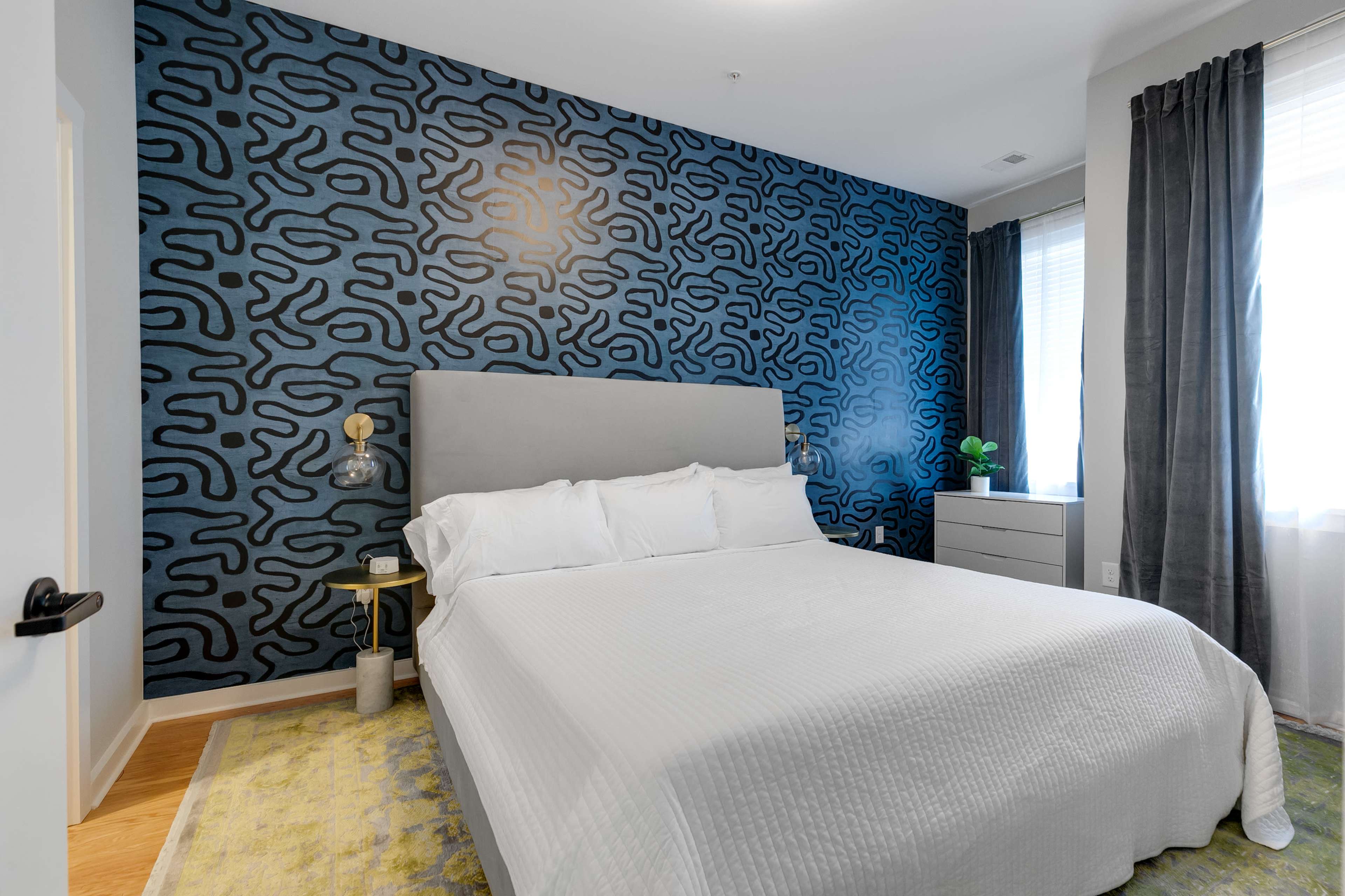 A bedroom with a gray upholstered bed against a blue patterned accent wall, complemented by white bedding and sheer curtains.