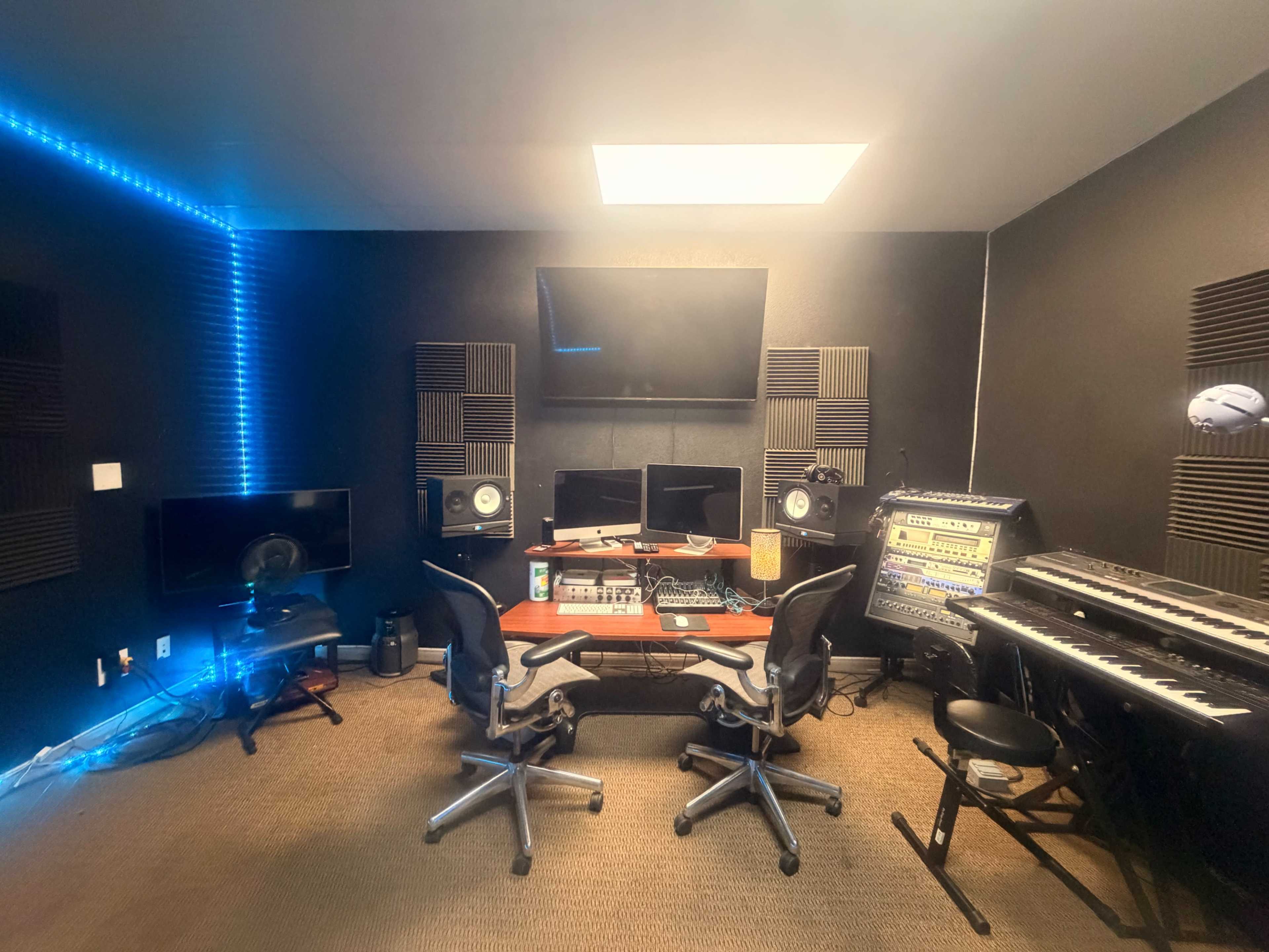 The image shows a music studio with a central wooden desk, computer monitors, audio equipment, and seating arranged around a dark, soundproofed room.