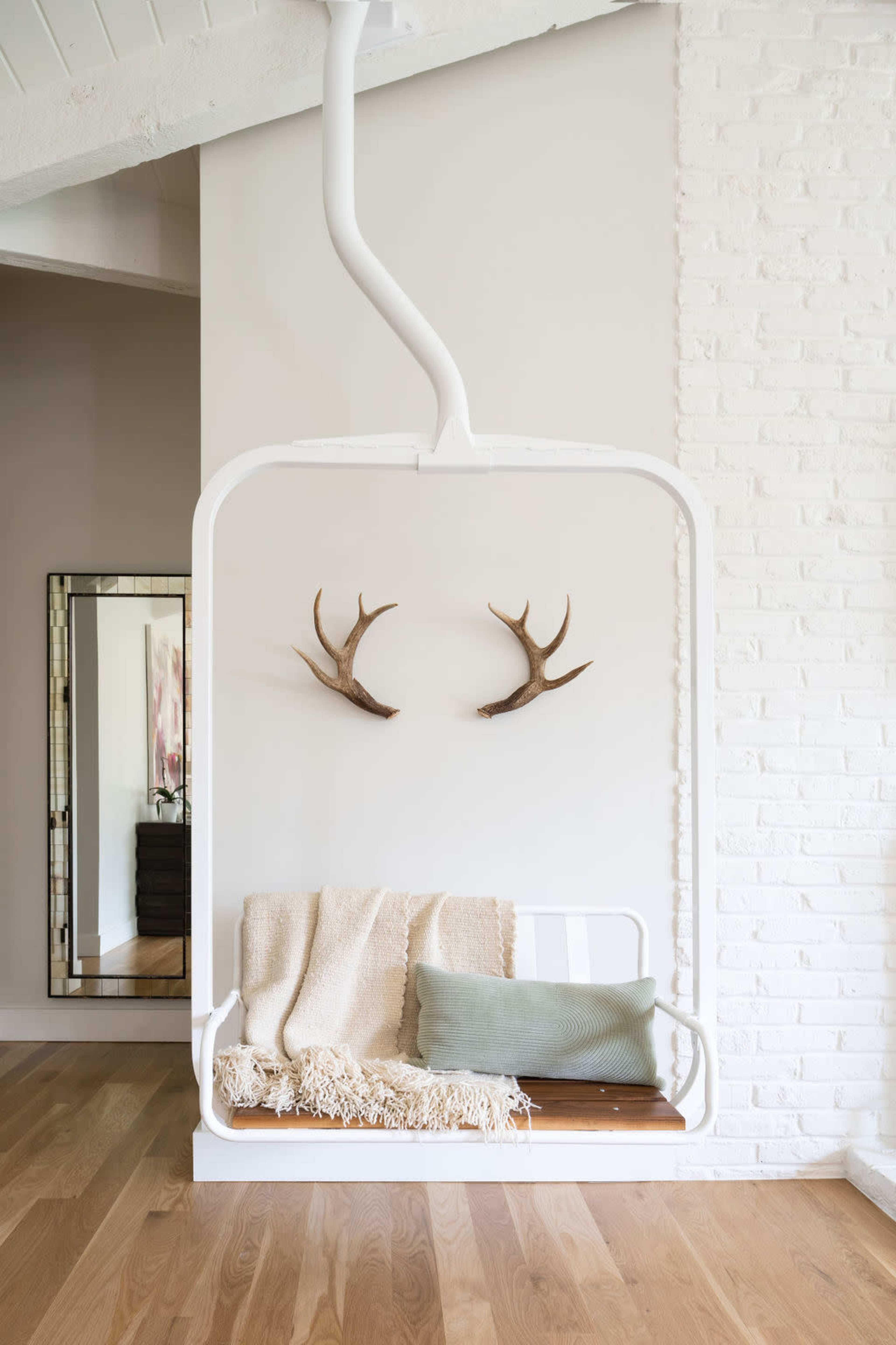 A white swing is mounted against a textured wall, adorned with deer antlers, a neutral blanket, and a green cushion, with a mirror reflecting the space.
