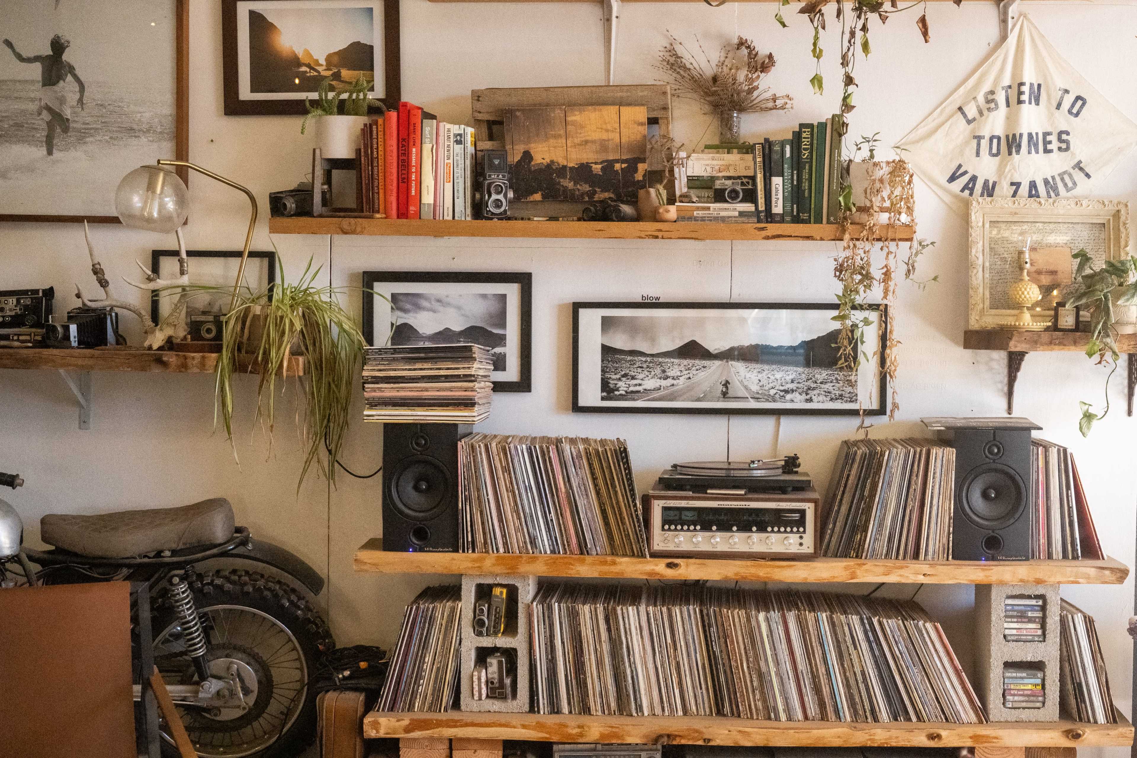 A collection of vinyl records is arranged on wooden shelves alongside books and framed photographs, with a vintage stereo system positioned in the center.