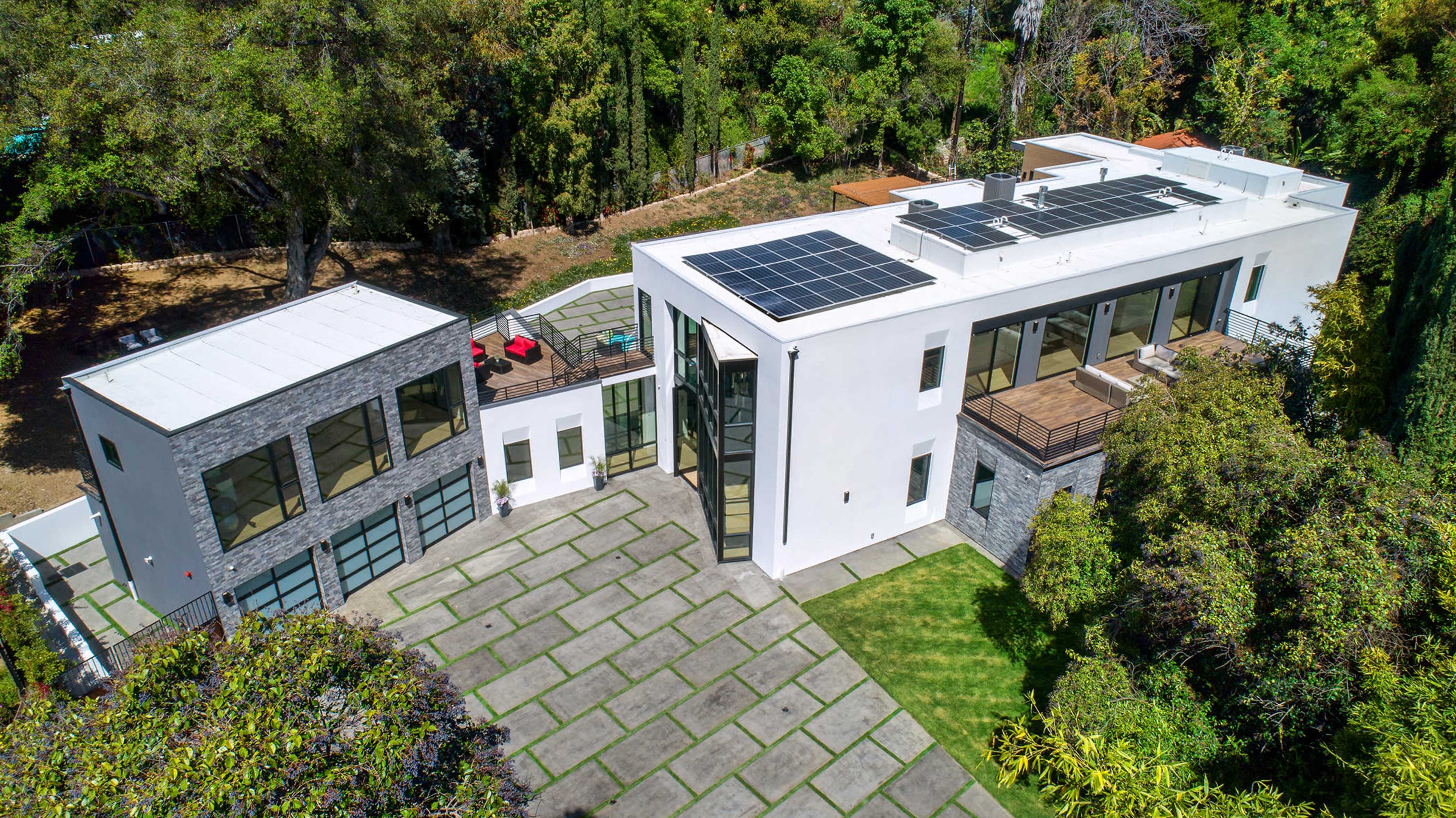 A modern two-story house with solar panels on the roof, surrounded by greenery and featuring a paved outdoor area.