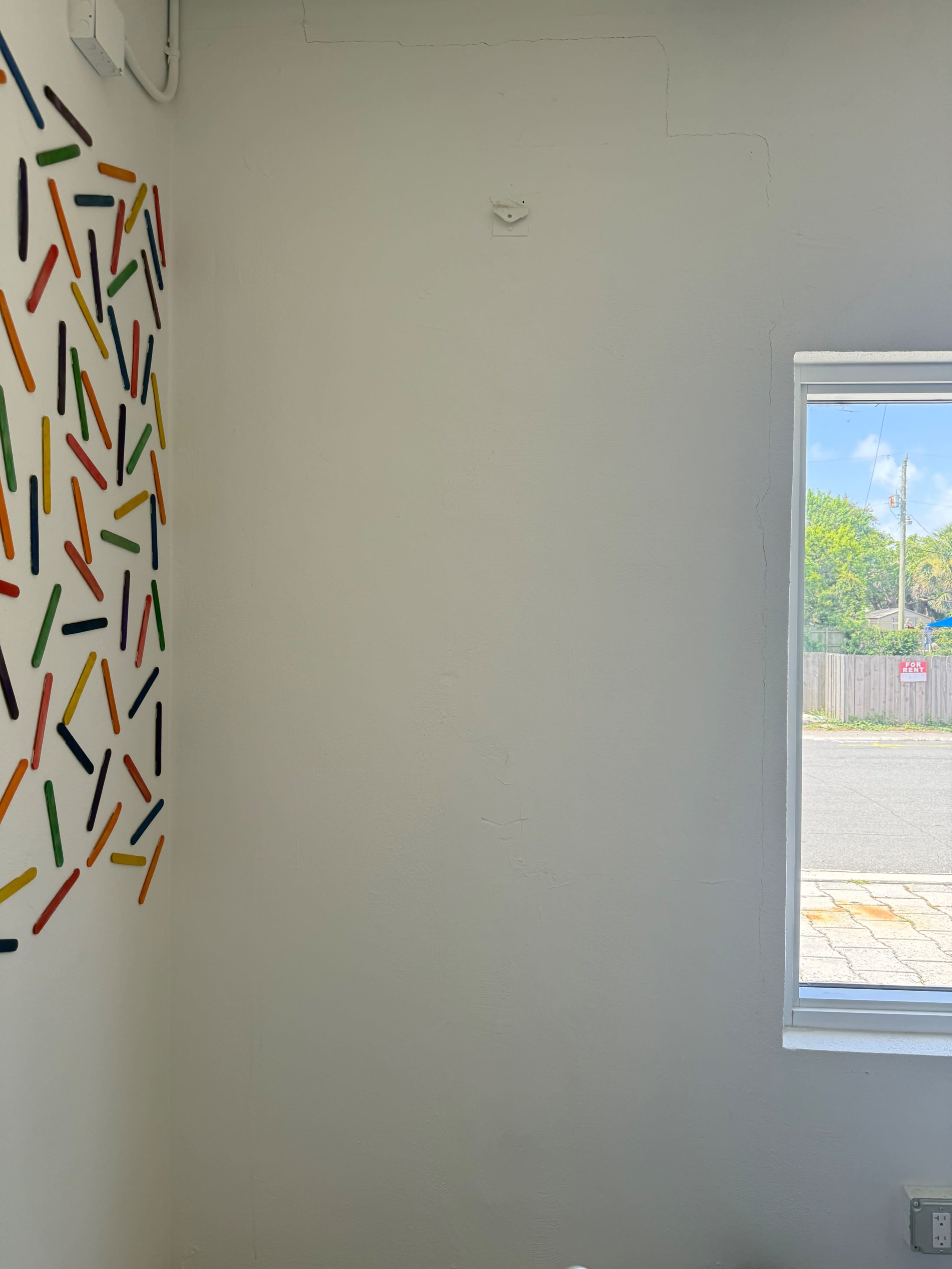 The image shows a corner of a room with a colorful arrangement of stick-like objects on one wall and a window on the adjacent wall, revealing a view of an outdoor scene.