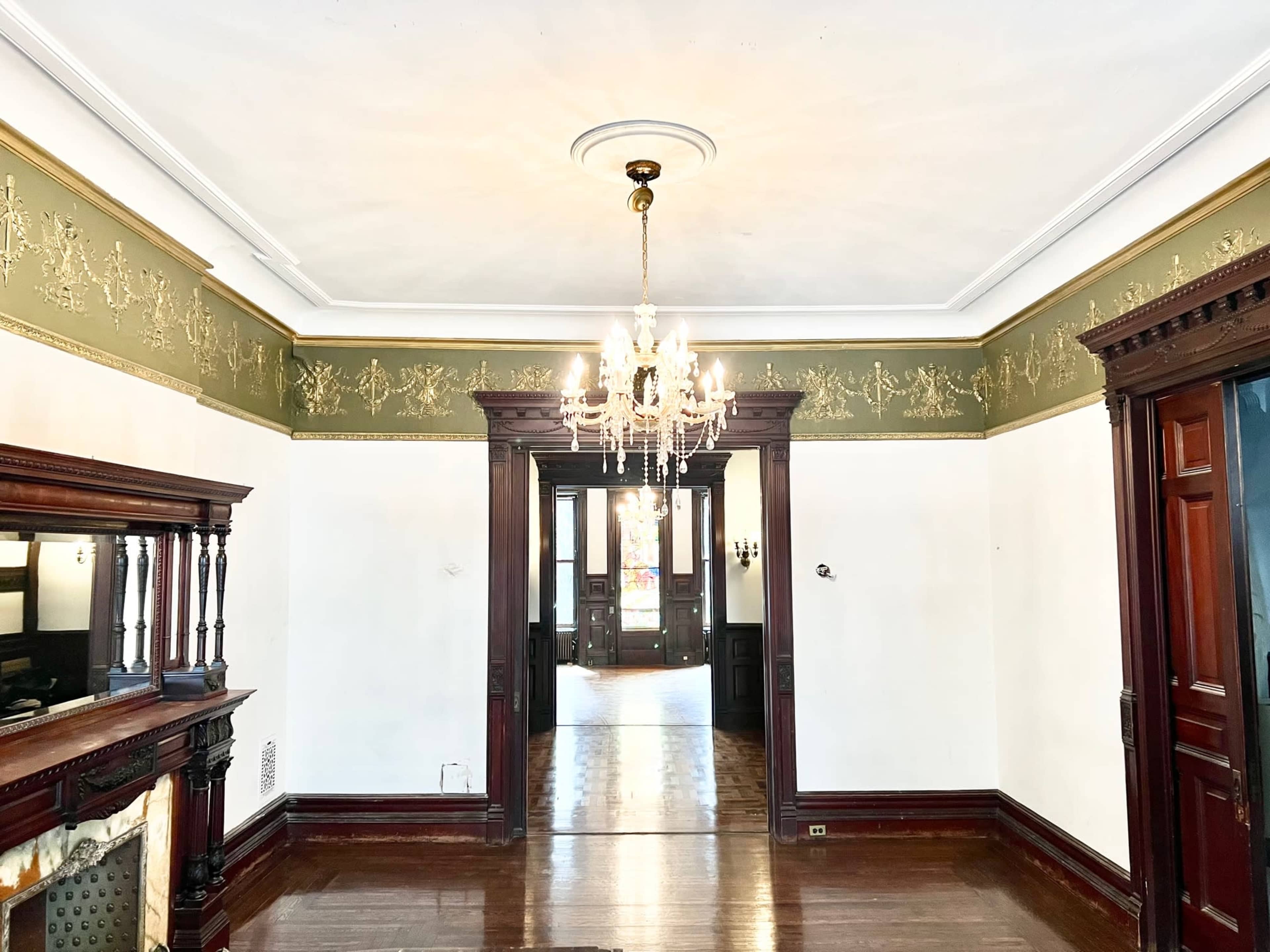 The image shows a spacious, empty room featuring ornate woodwork, a chandelier, and a decorative green wall border.