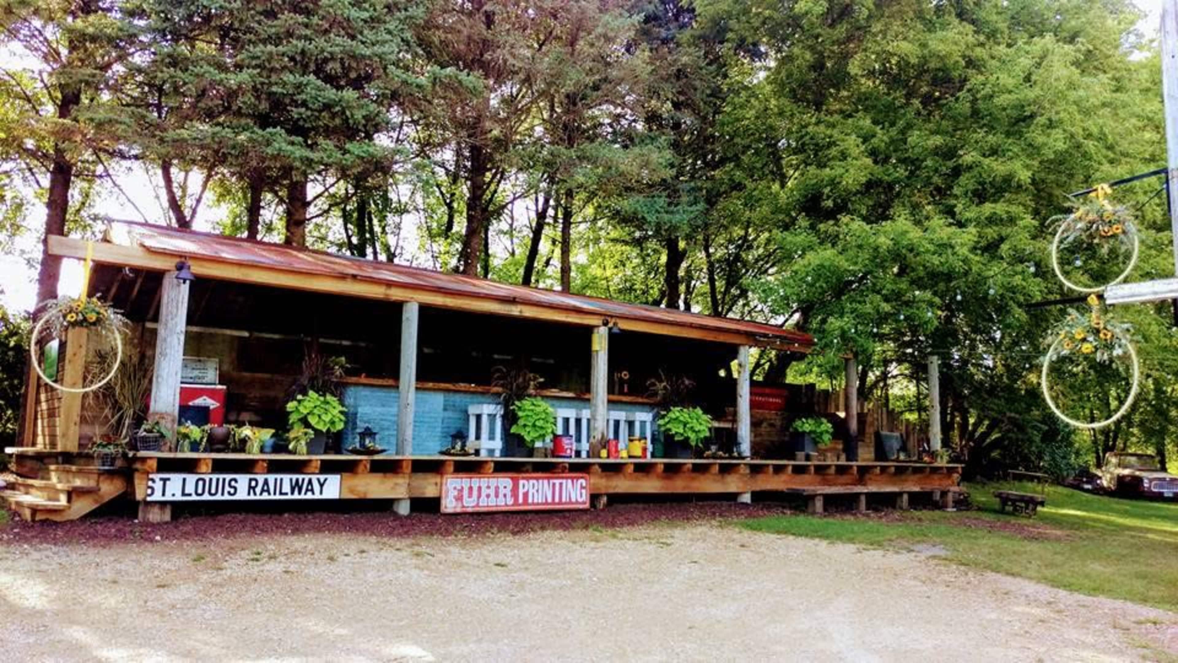 The image shows a wooden structure resembling a railway station, adorned with potted plants and surrounded by trees.