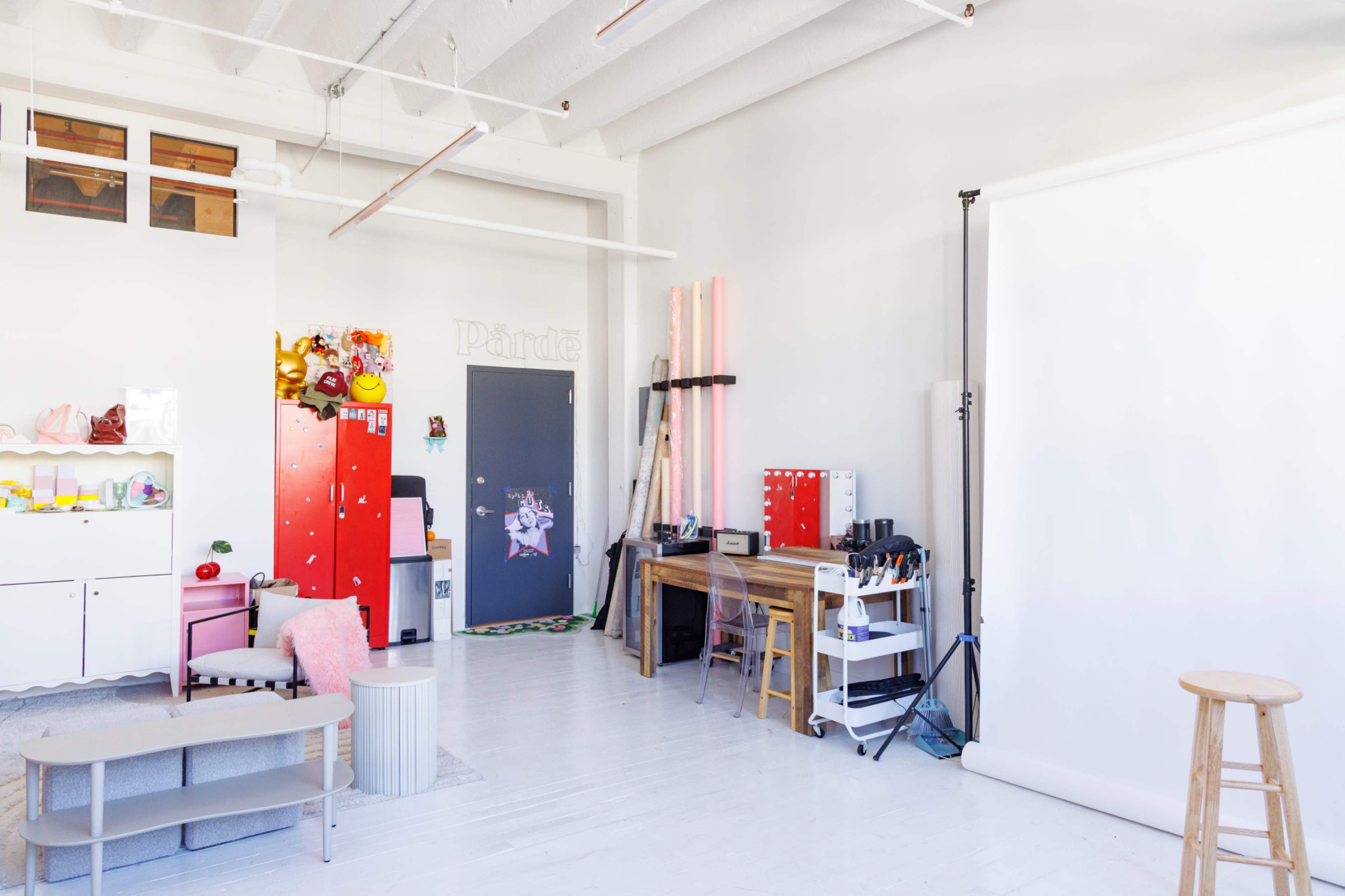 The image shows a bright, airy studio space with a white backdrop, colorful decorations, a red fridge, and a wooden work table surrounded by various equipment and props.