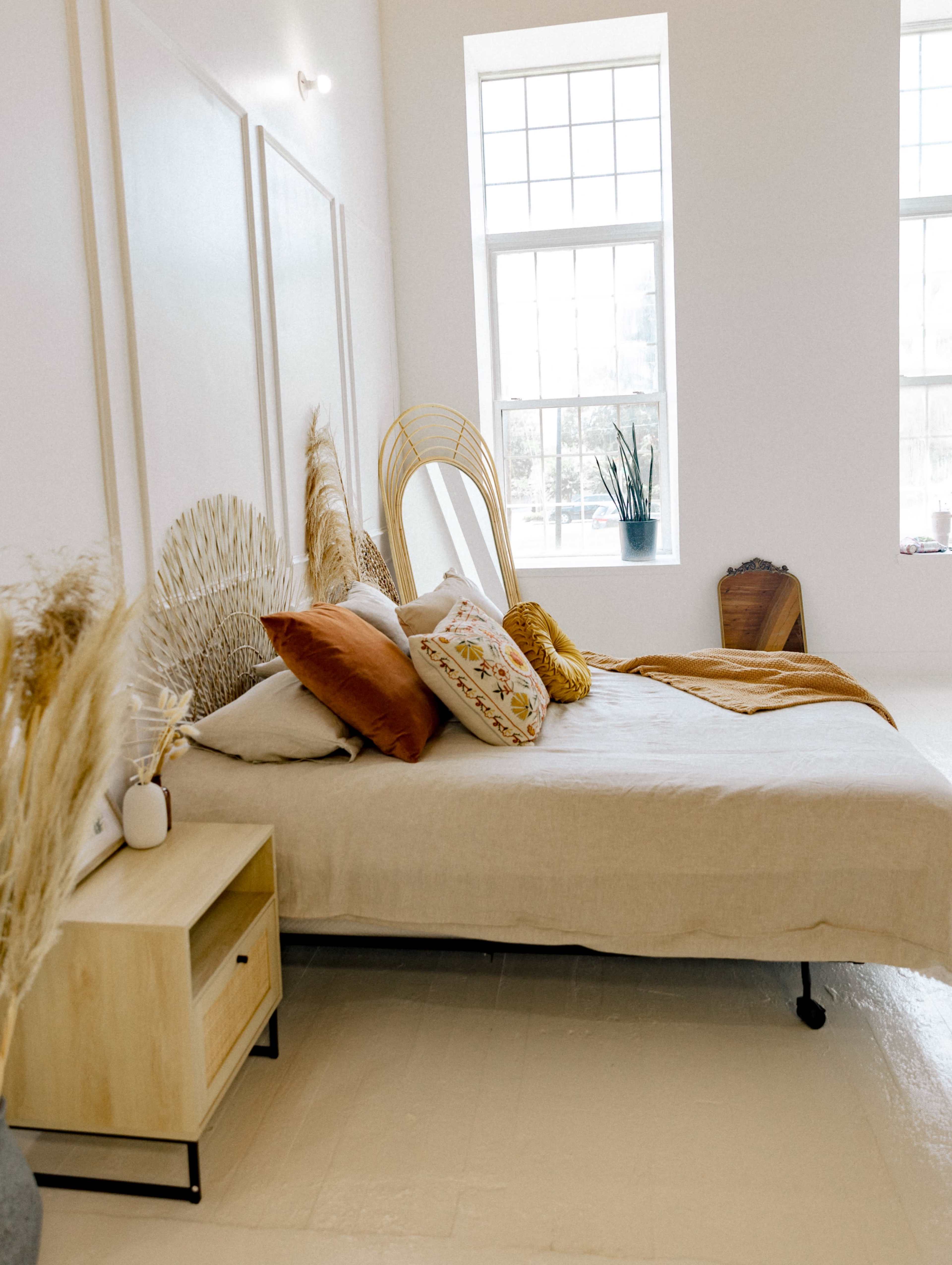 A light-filled bedroom features a bed adorned with decorative pillows and a woven headboard, accompanied by a bedside table and large windows.