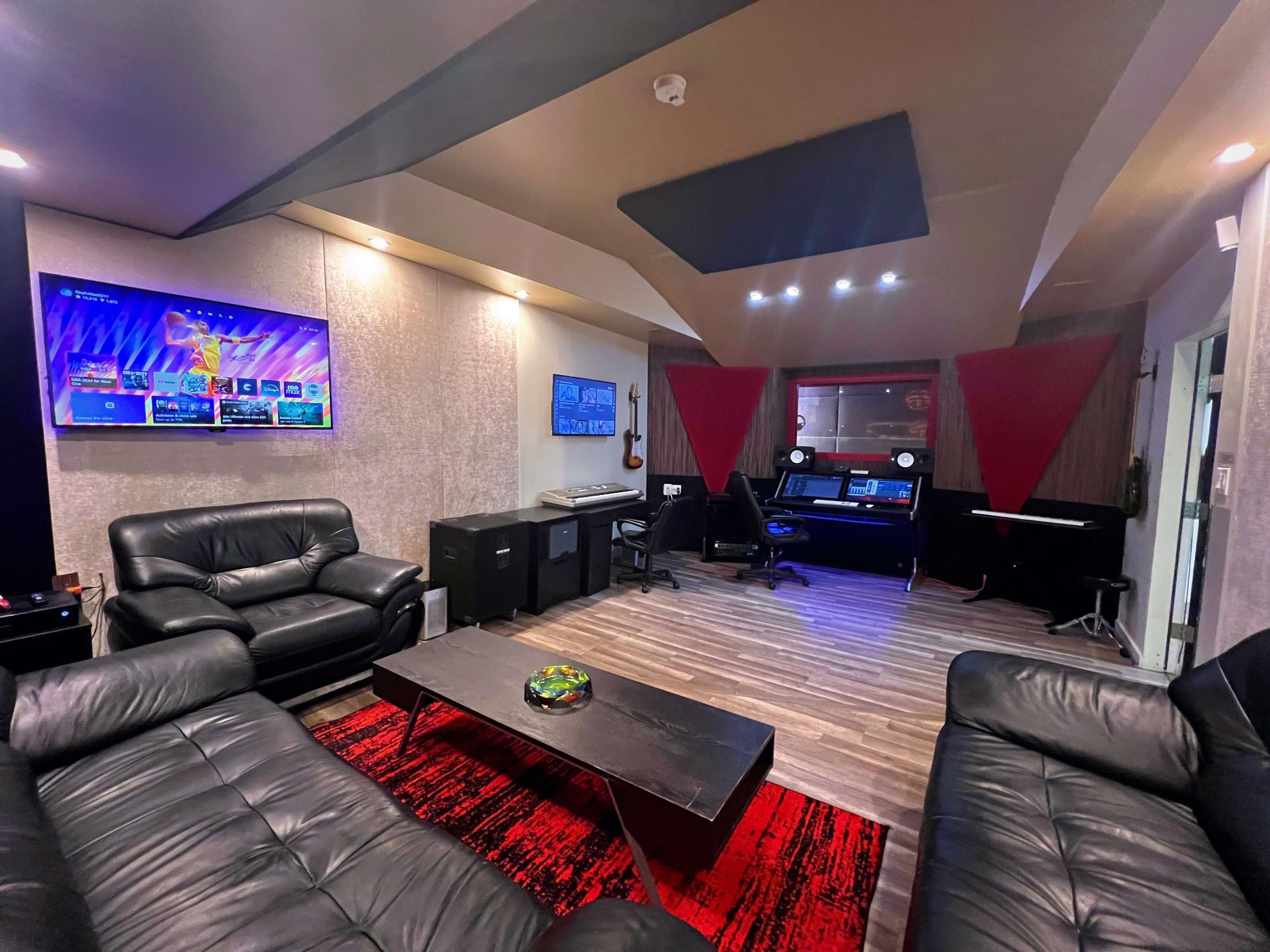 A modern recording studio with black leather sofas, a coffee table, and audio equipment arranged against a backdrop of wood and gray walls.