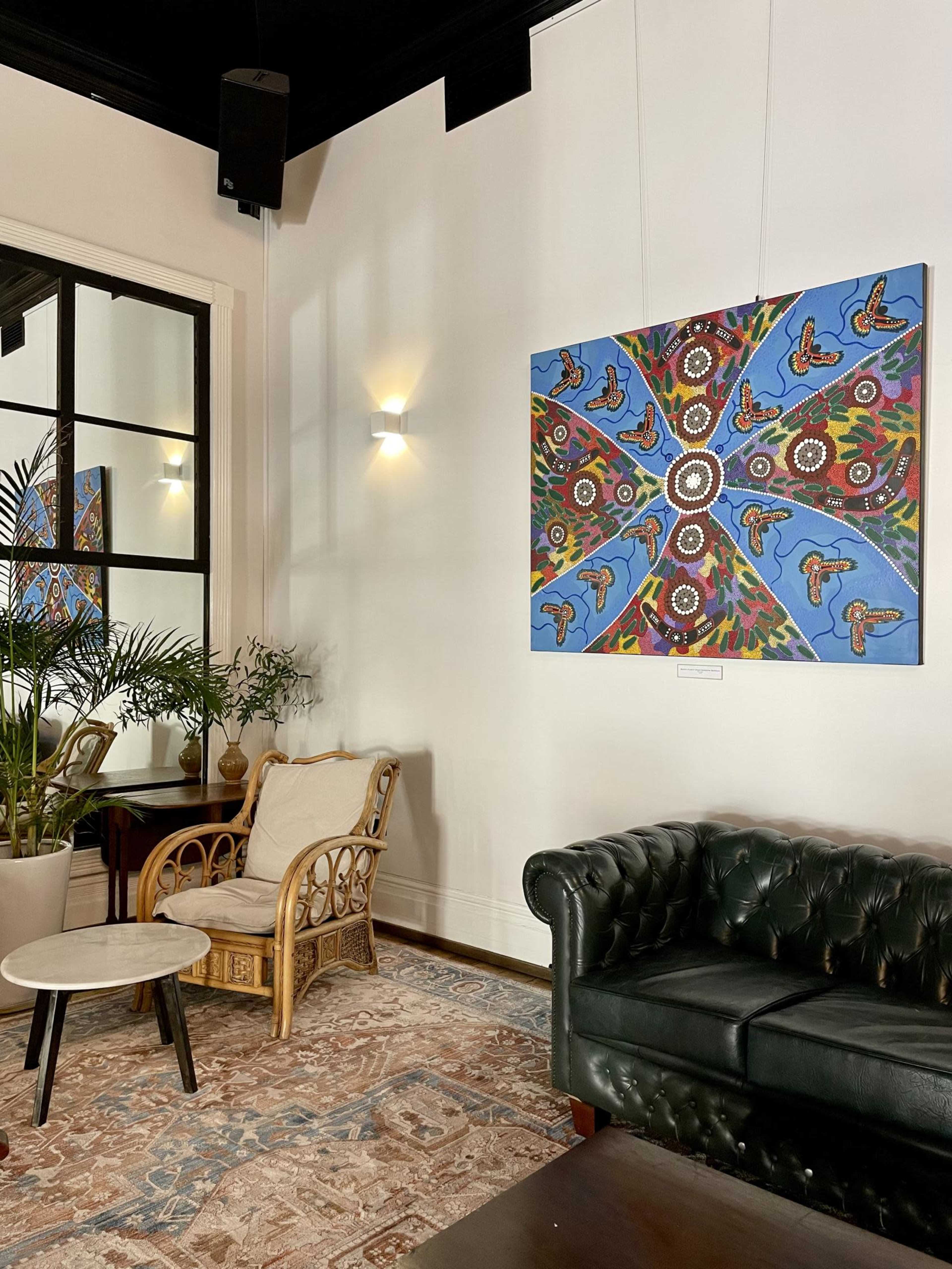 The image shows a cozy indoor space featuring a green leather sofa, a rattan chair, a small round table, and a colorful artwork hung on the wall.