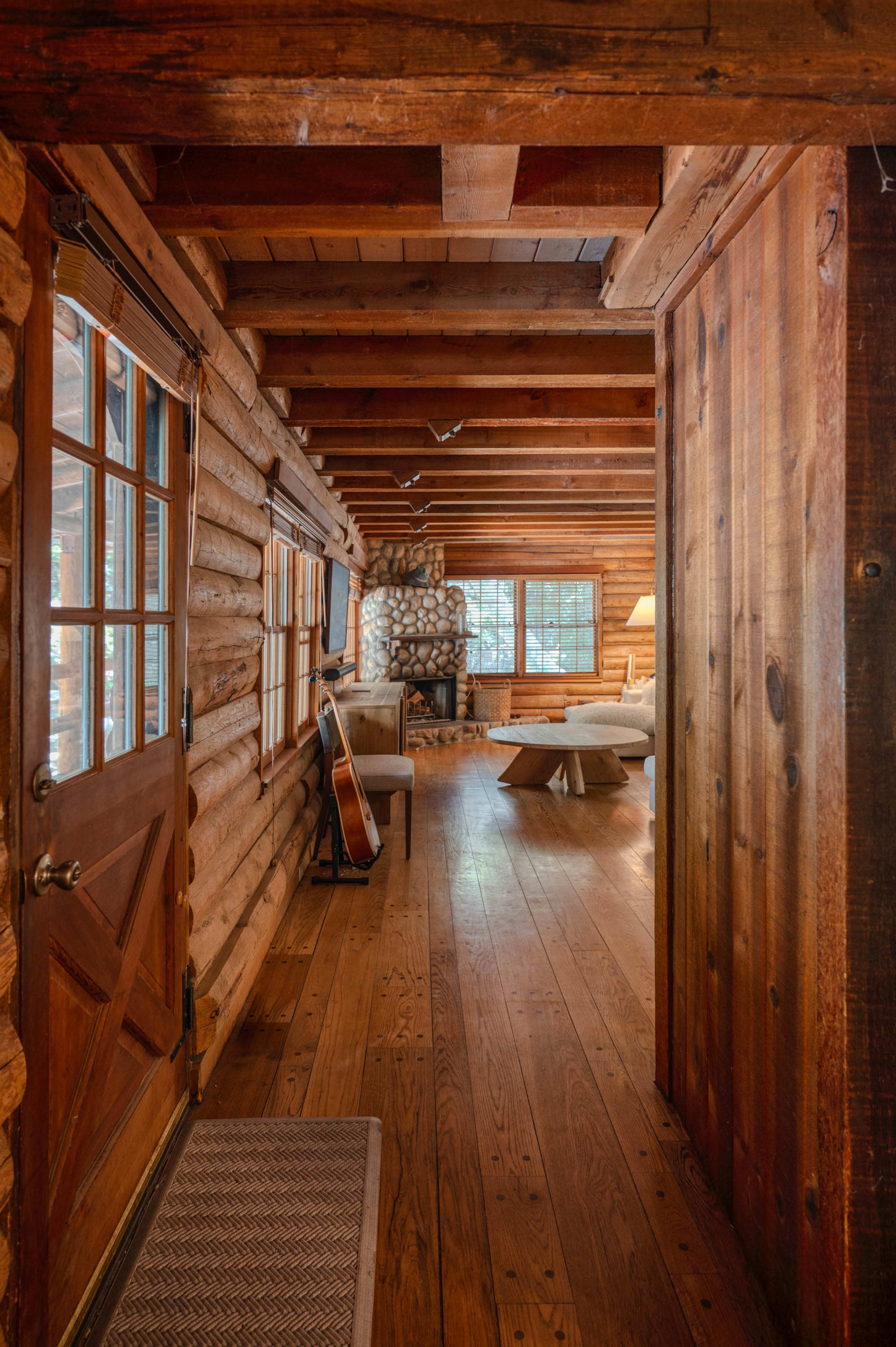 The image depicts a log cabin interior featuring a hallway that leads to a living area with wooden floors and a stone fireplace.