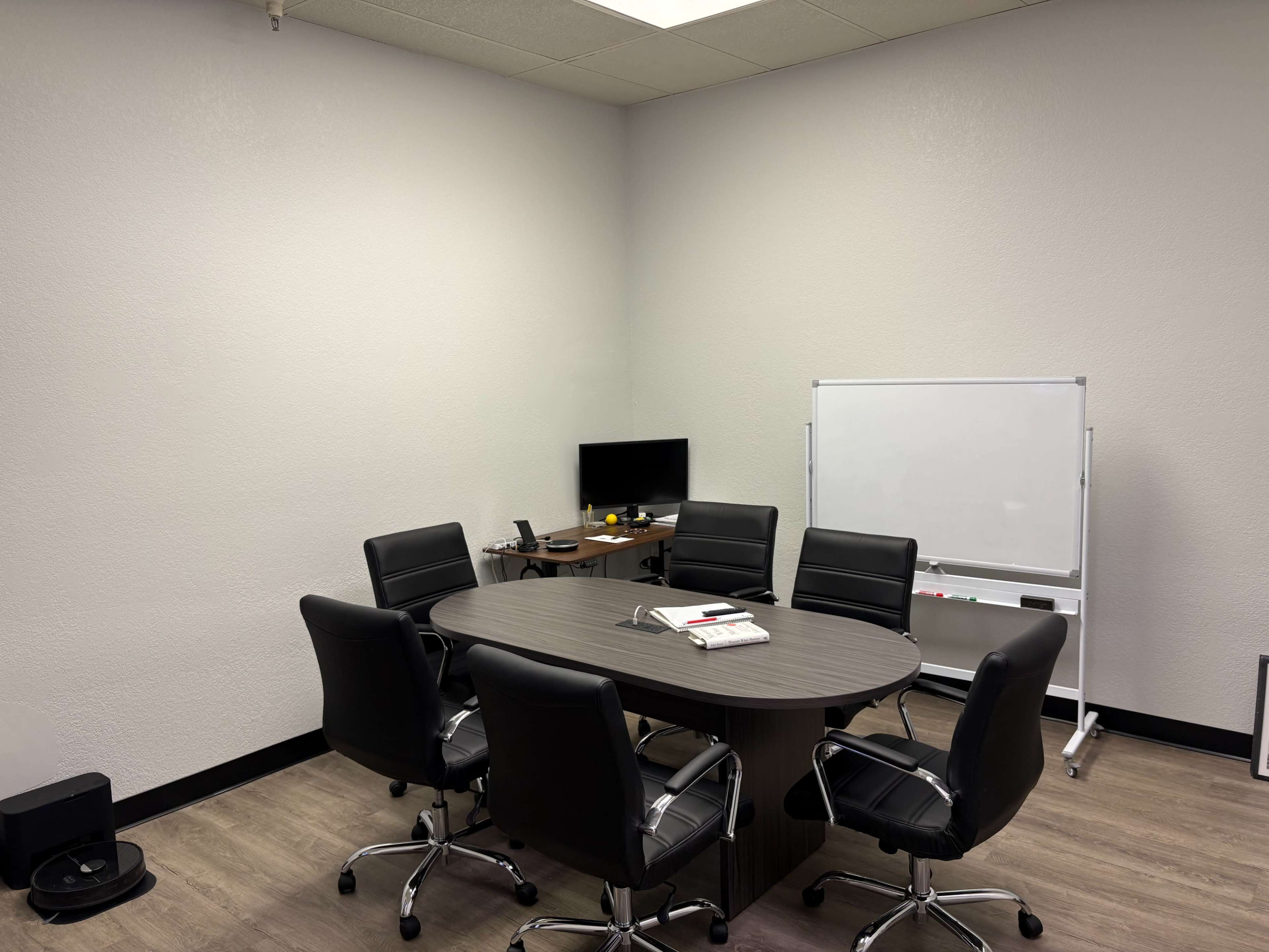 The image shows a small conference room with a round table surrounded by six black chairs, a computer monitor on a desk, and a whiteboard on a stand.