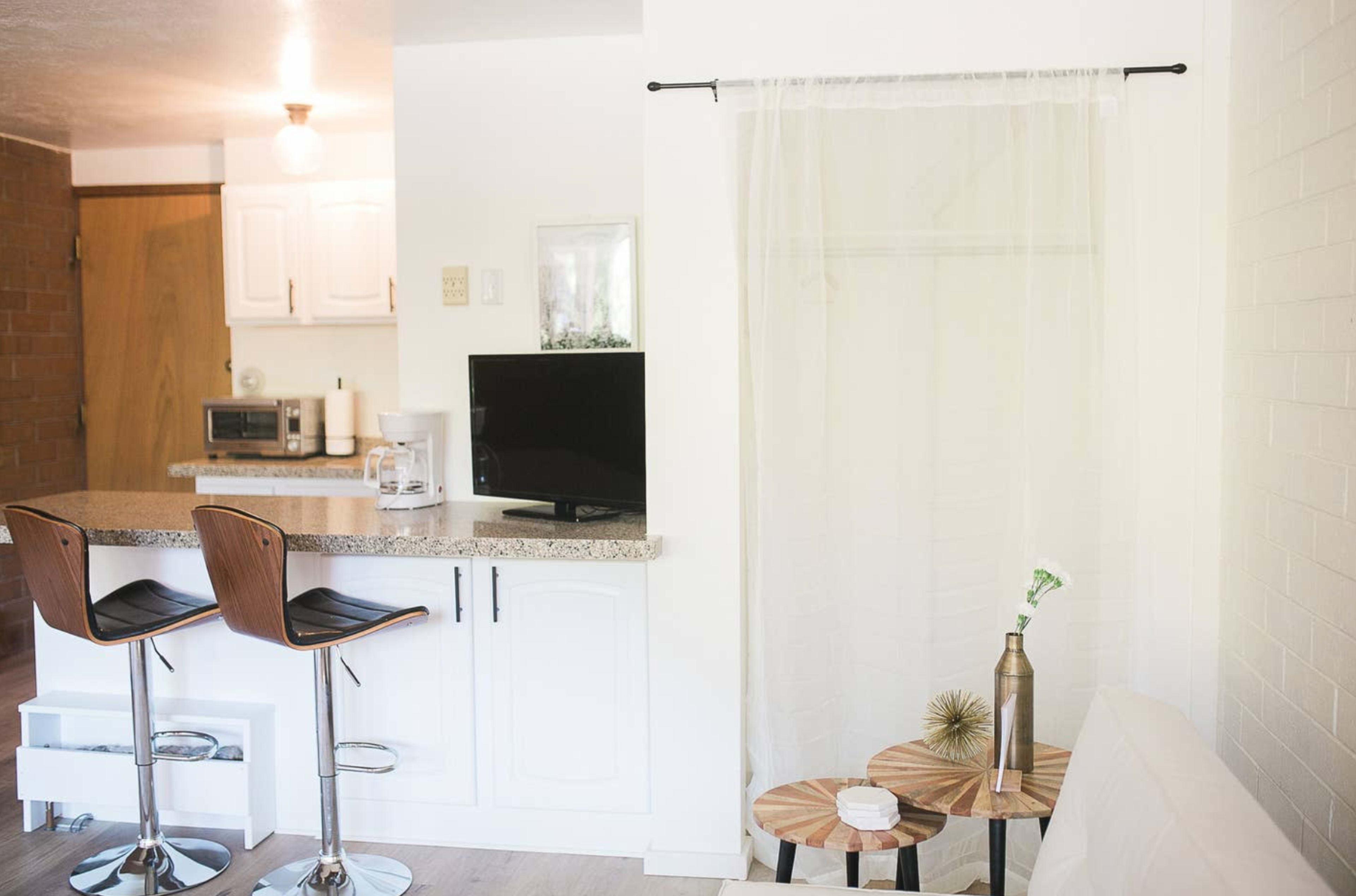 A bright, modern living space with a kitchenette featuring a bar counter and two stools, a small TV on the counter, and a curtain separating the area from a seating space.