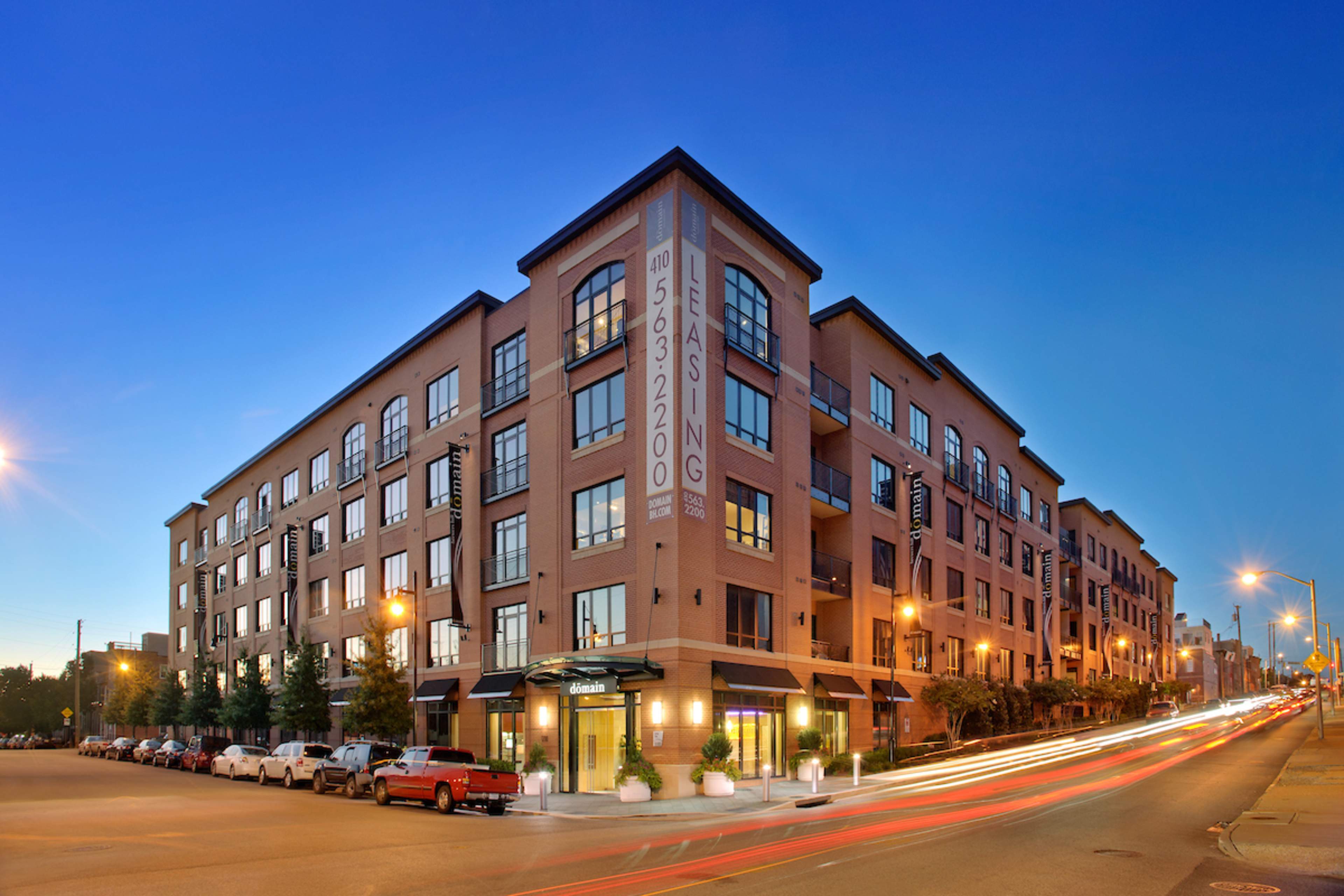 A four-story brick apartment building with leasing signage, illuminated by streetlights, at dusk with moving car lights creating a streak effect in the foreground.