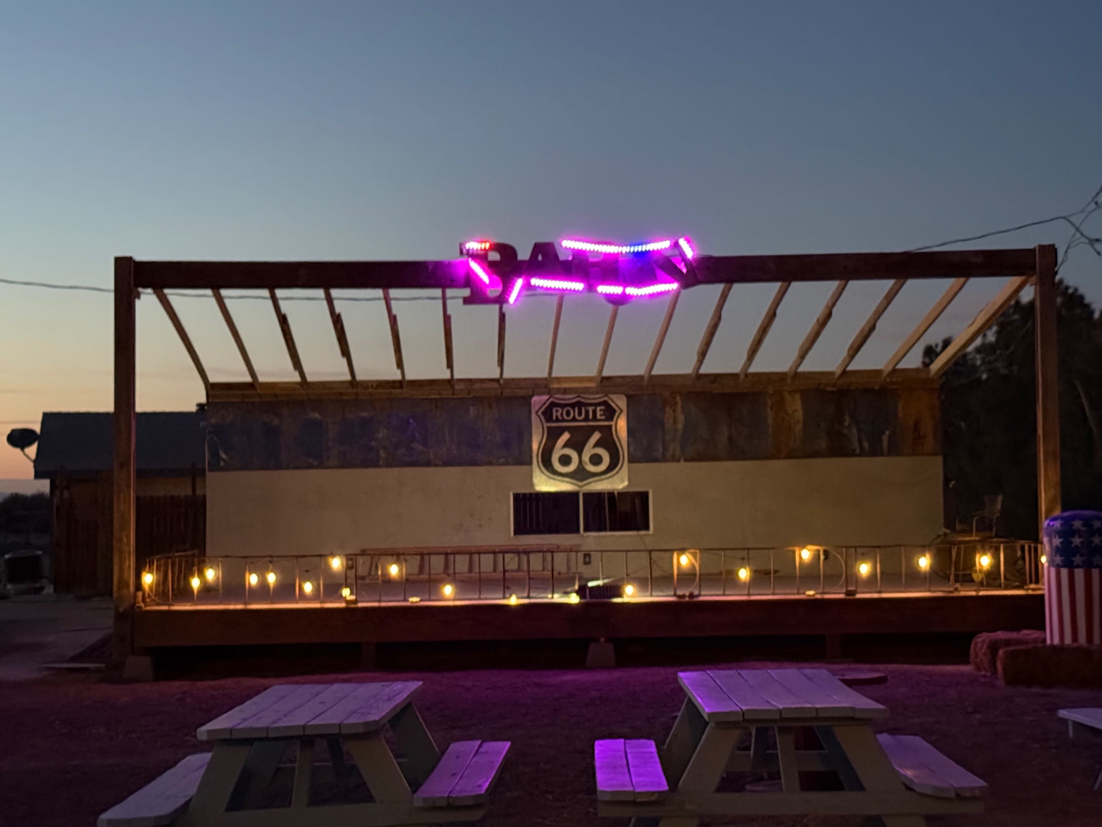 The image shows a wooden stage with a Route 66 sign and neon lights above it, illuminated by small lights at dusk.