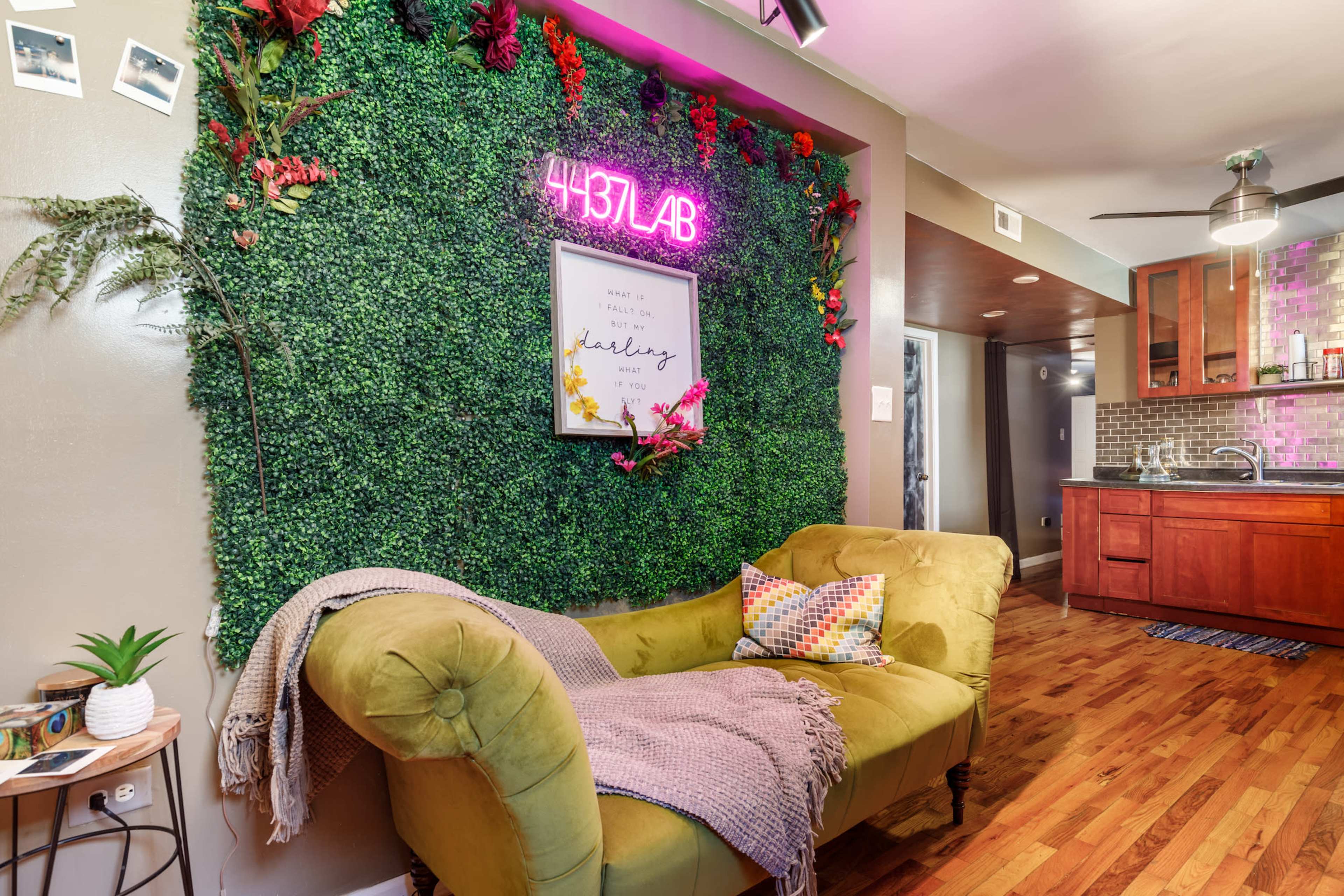 A cozy living space featuring a vibrant green wall adorned with faux plants and a neon sign, alongside a vintage-style yellow couch and a modern kitchenette in the background.