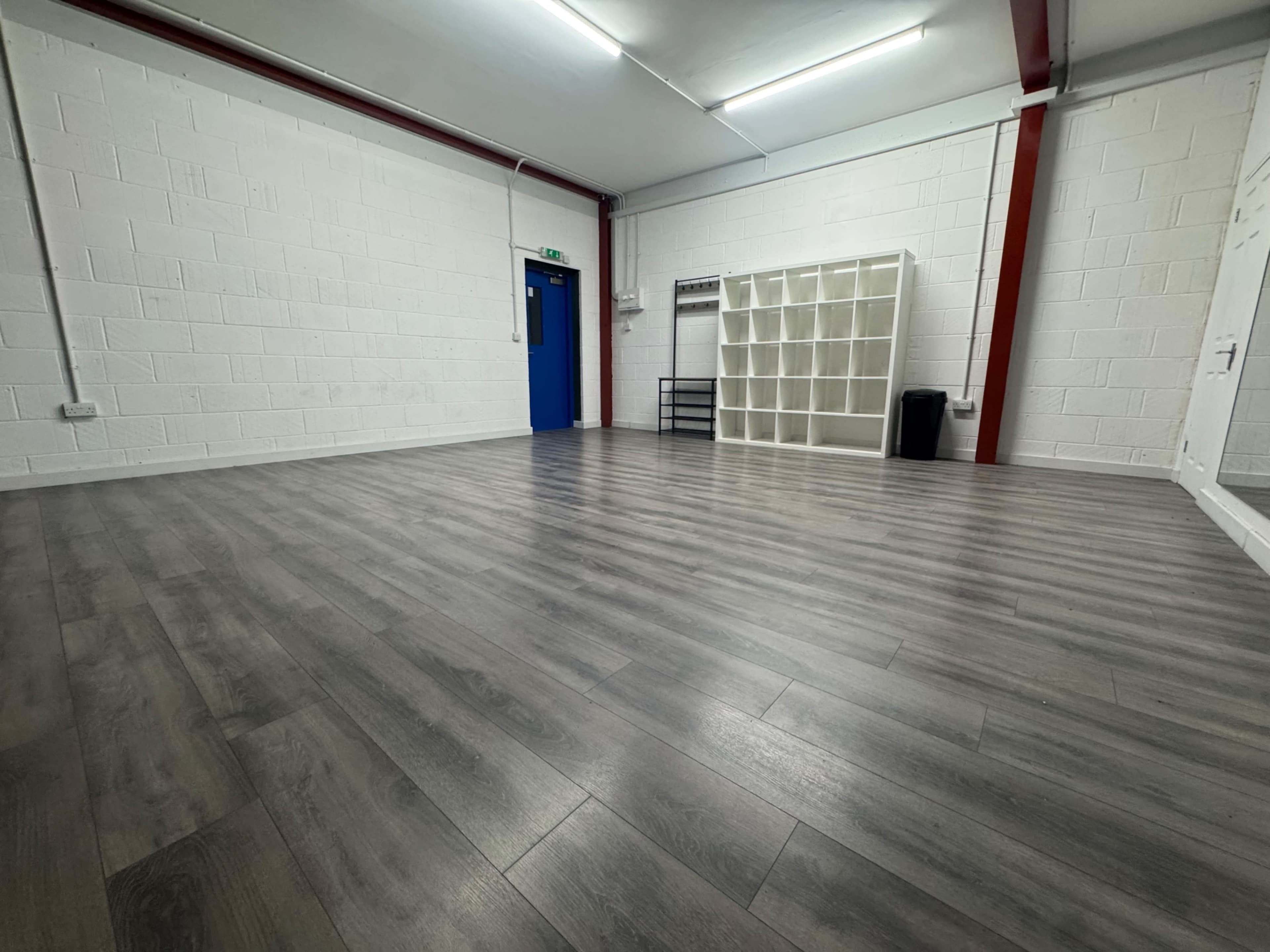 The image shows a spacious, empty room with grey laminate flooring, white walls, a blue door, and minimal furniture, including a shelving unit.
