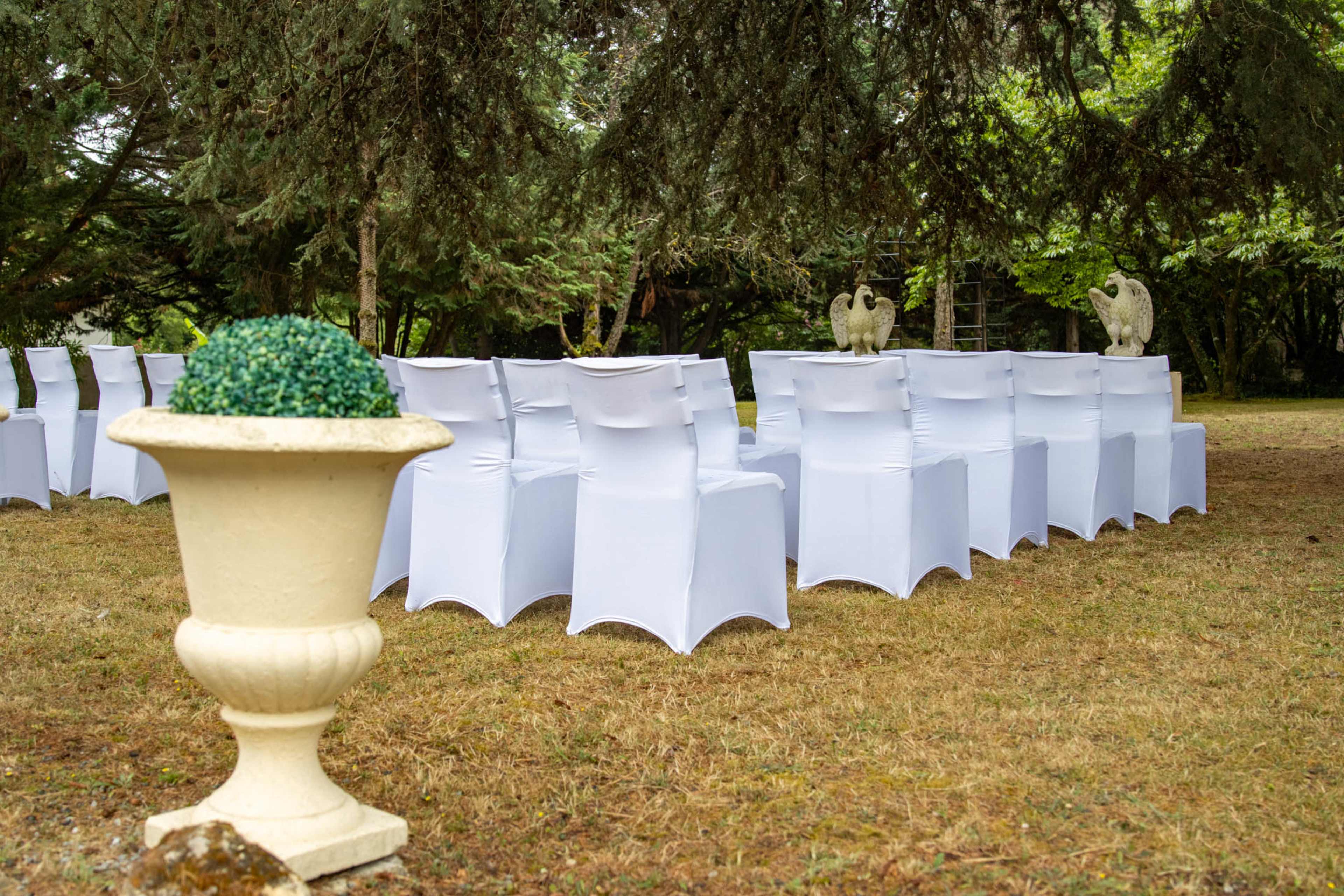 Chairs with white covers are arranged in rows on a grassy area, with ornamental statues and a decorative planter in the foreground.