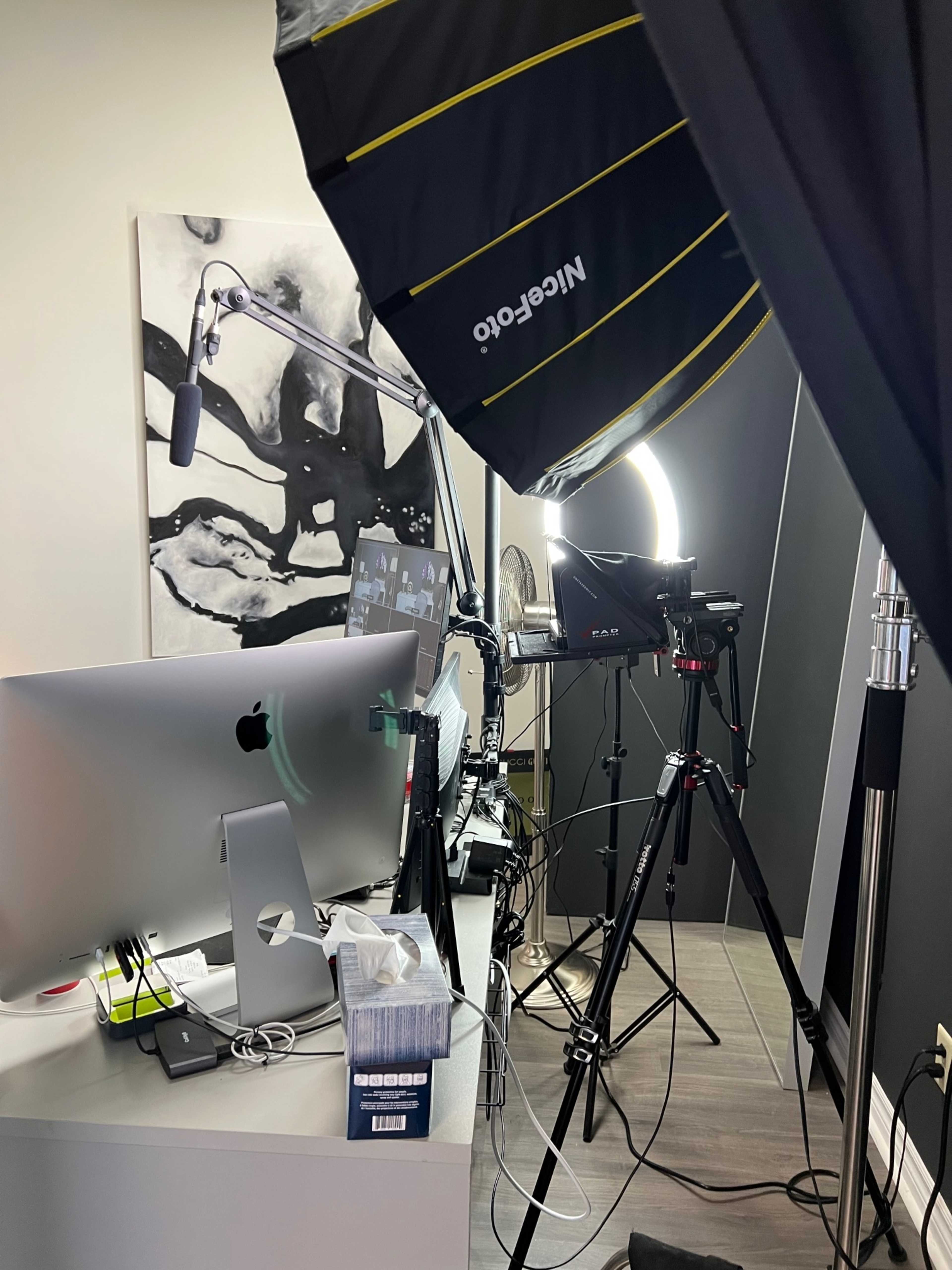 The image shows a workspace setup with a computer, camera equipment, lighting umbrellas, and various office supplies arranged in a room.