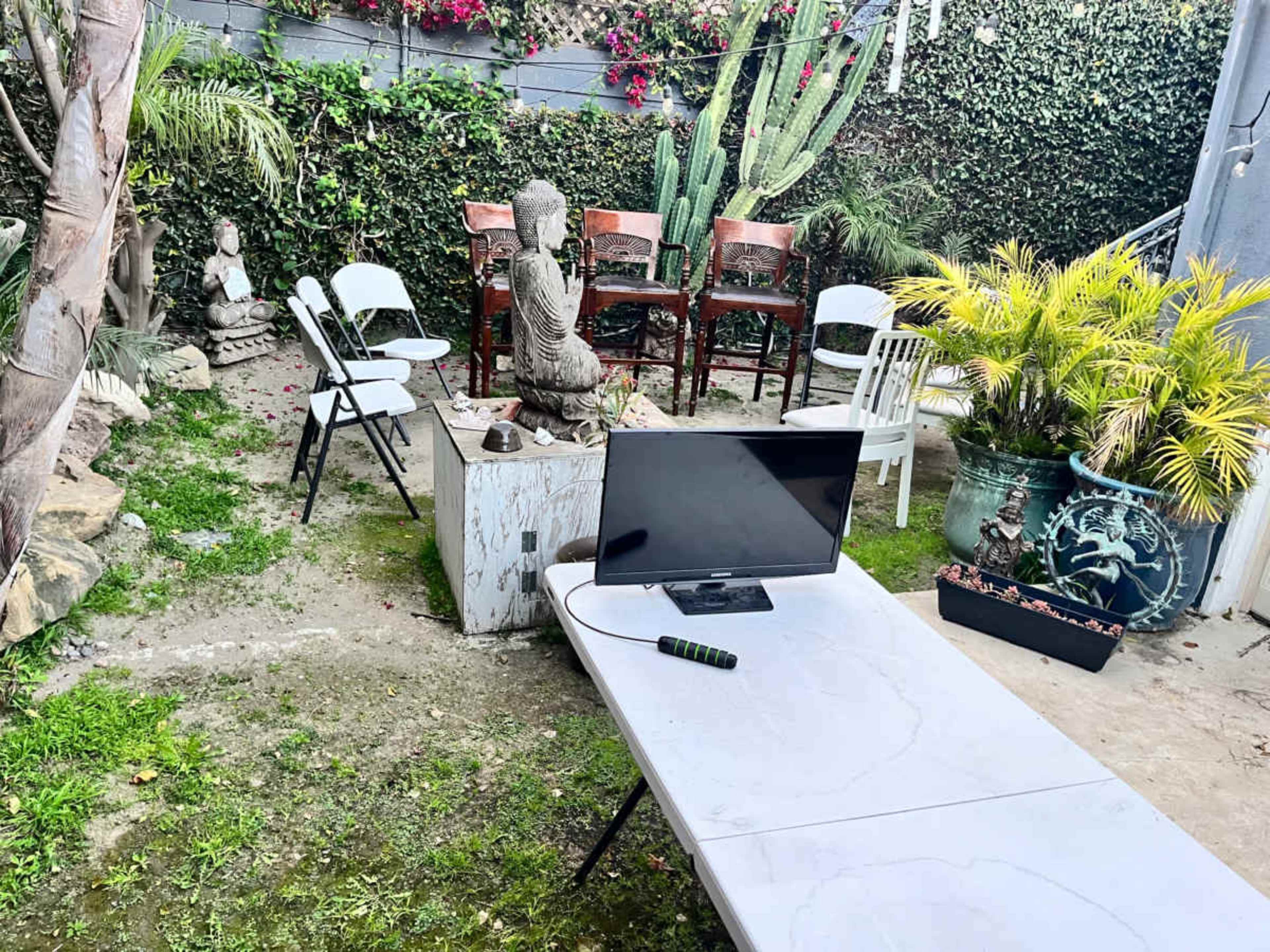 A small outdoor space features a table facing a television, surrounded by chairs, sculptures, and plants.