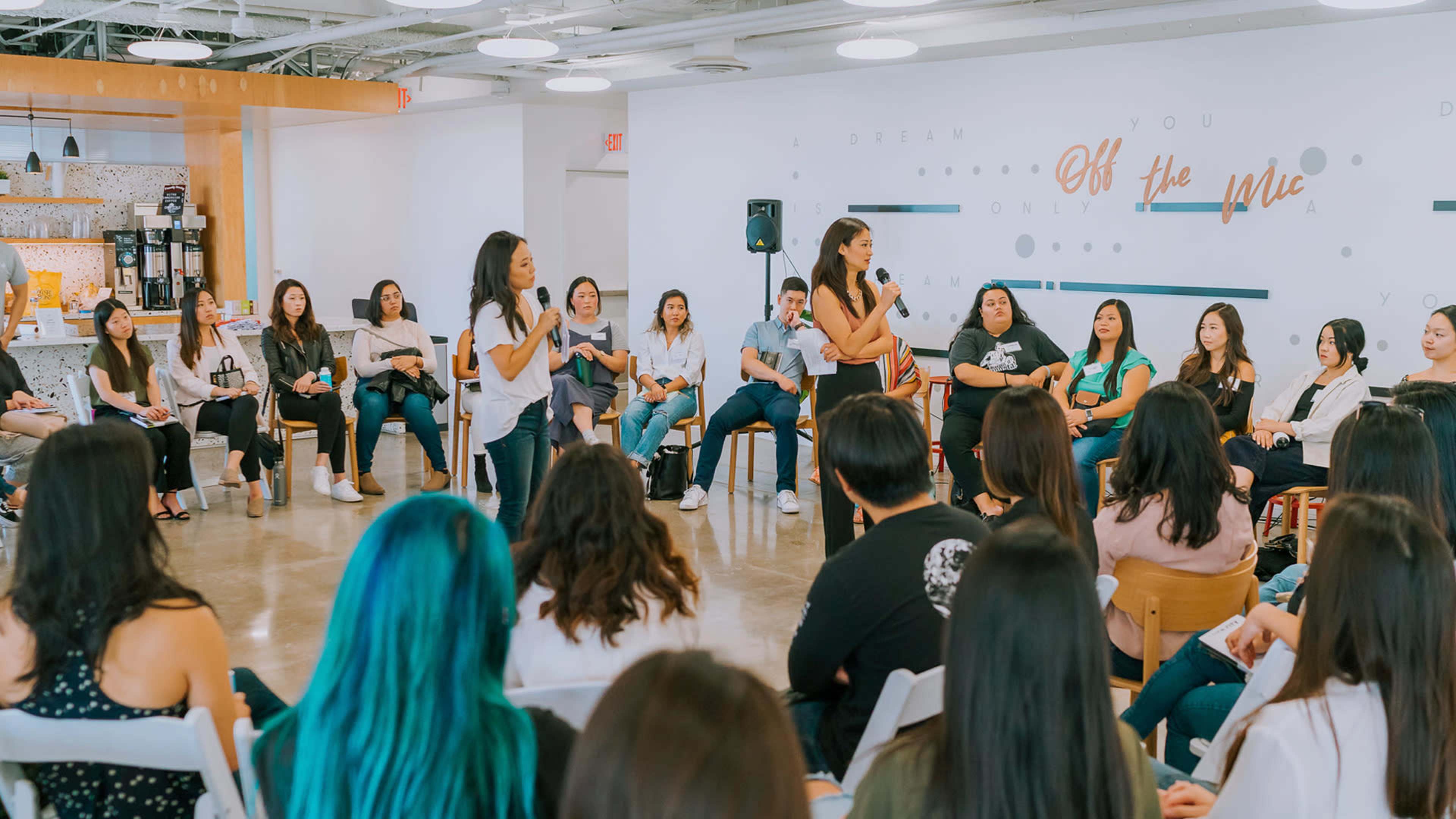 A group of people sits in a circle while two individuals speak in the center of a well-lit room.