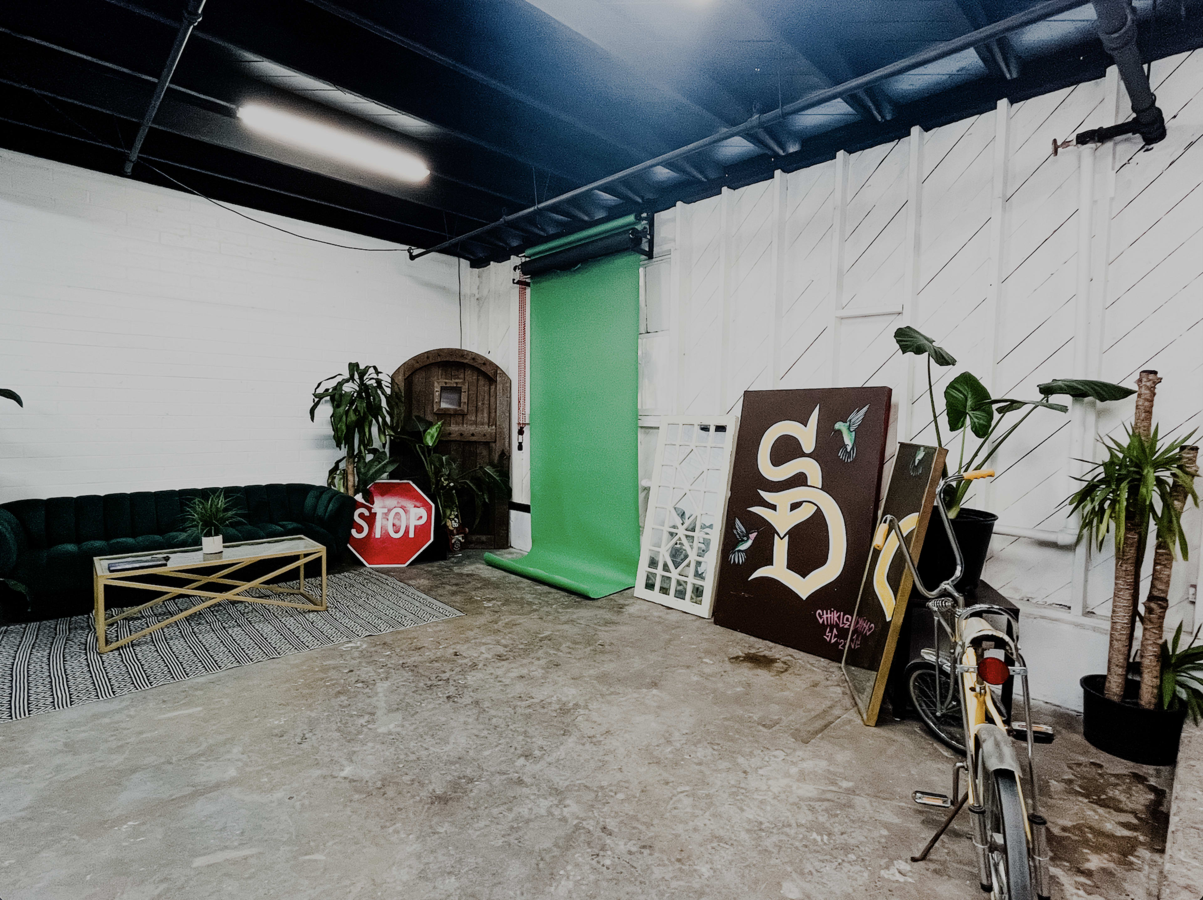The image shows a spacious studio with a green backdrop, a dark green couch, a stop sign, and various plants scattered around.