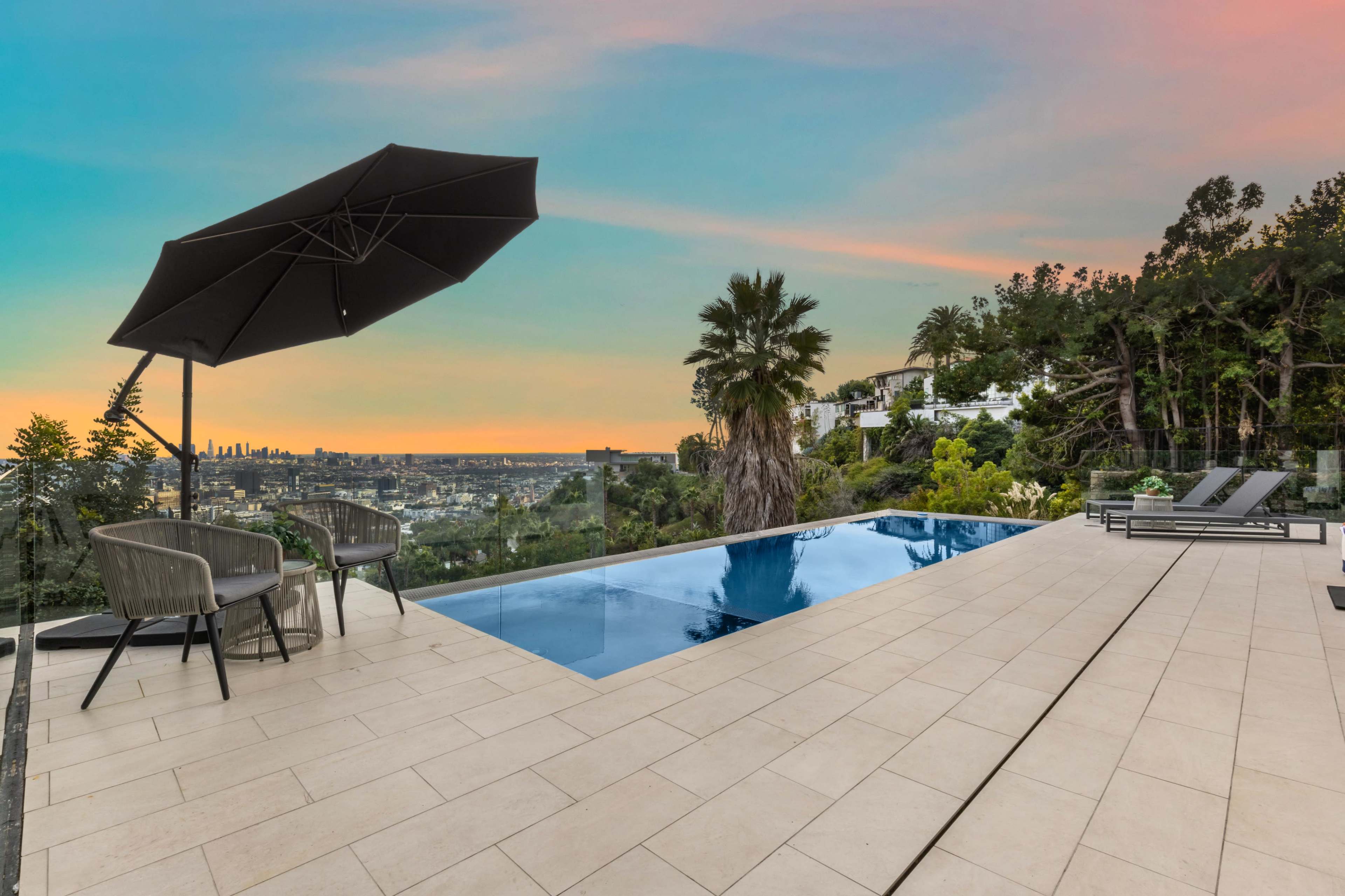 The image shows a modern terrace with a swimming pool, two chairs under an umbrella, and a panoramic view of a city against a colorful sunset sky.