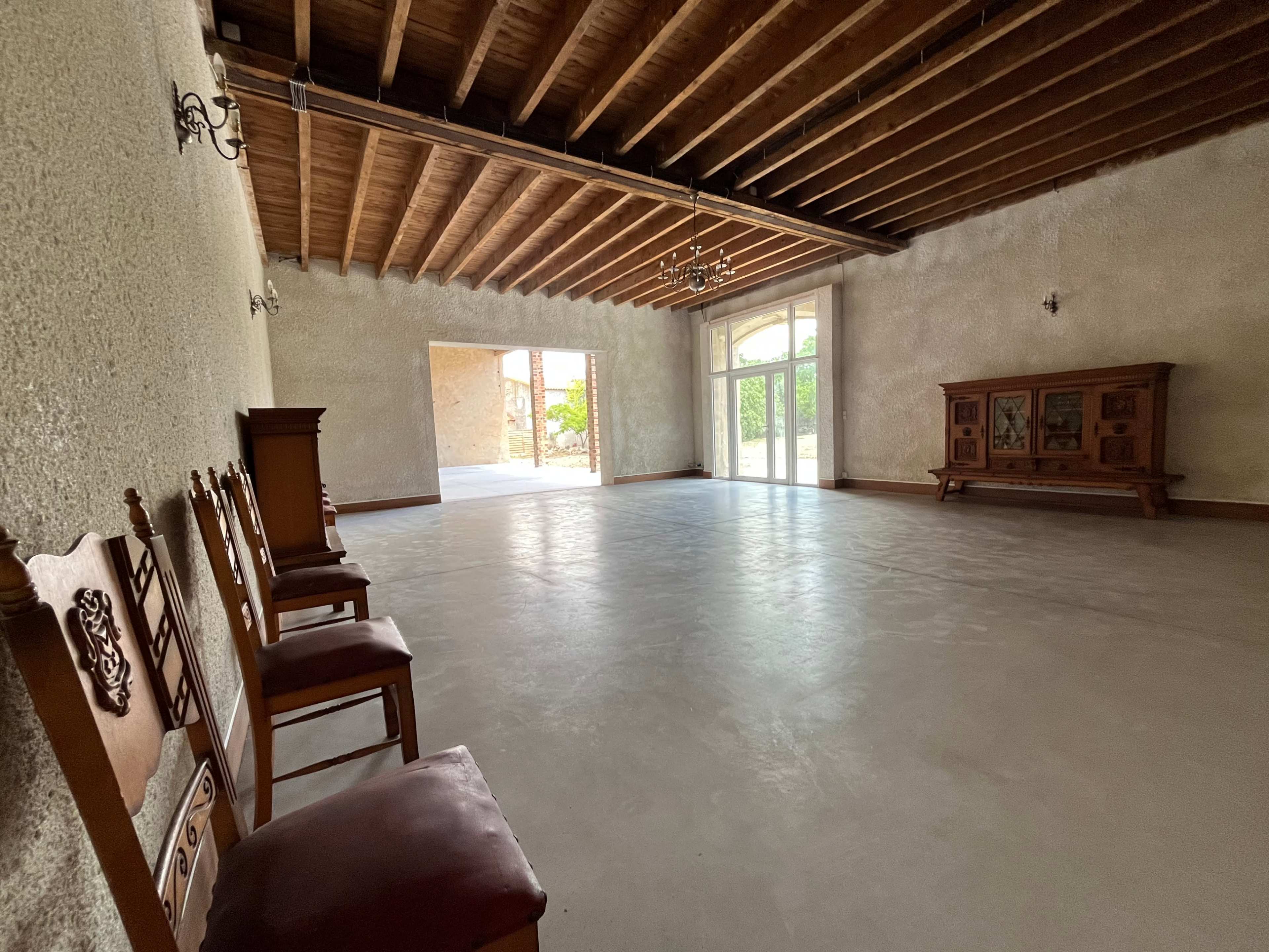 The image shows an empty room with wooden beams on the ceiling, a few wooden chairs aligned against one wall, and a large window opening to an outdoor area.