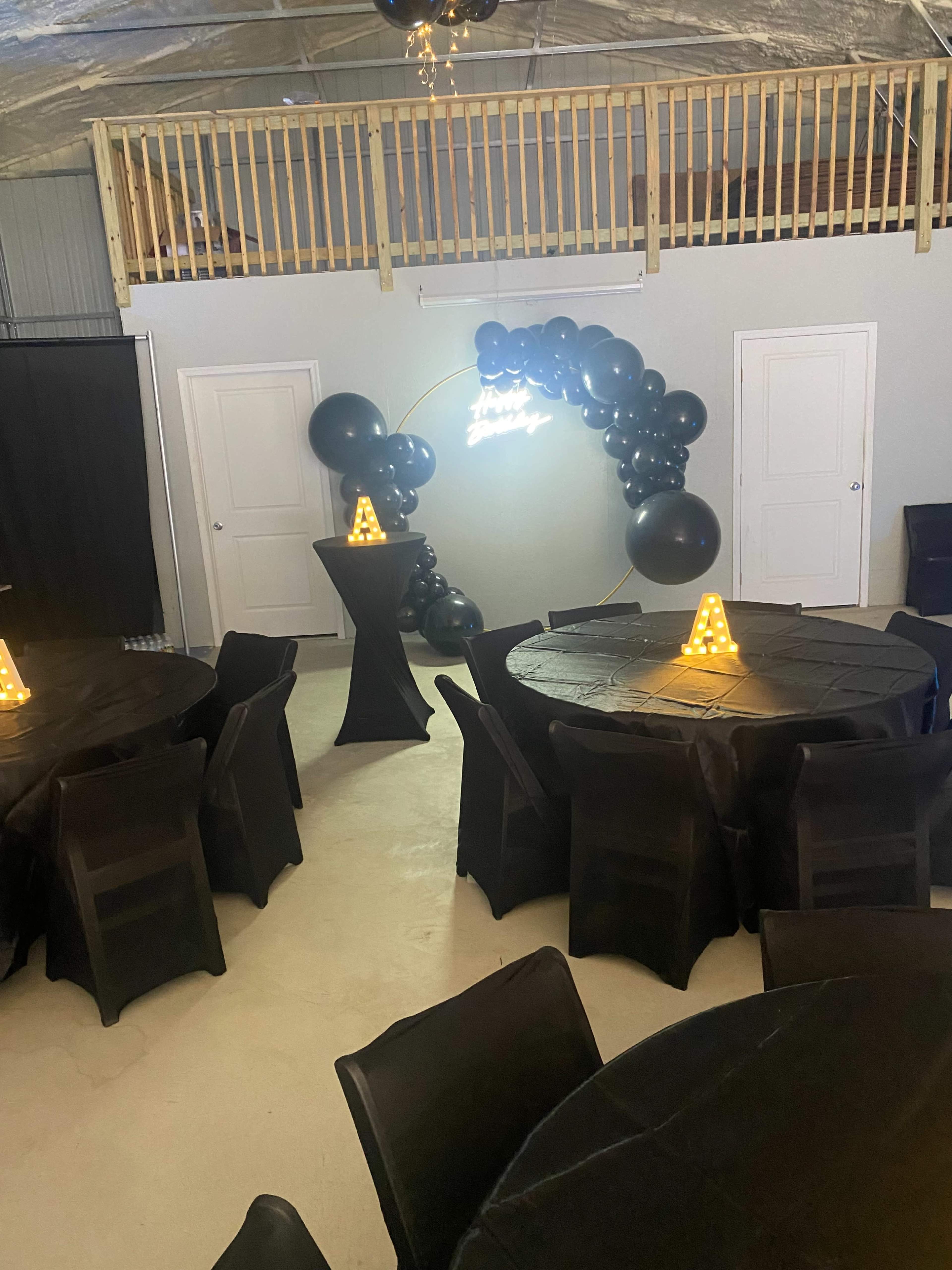 The image shows a spacious event space featuring black tables with tablecloths, illuminated centerpieces, and a decorative arch made of black balloons.