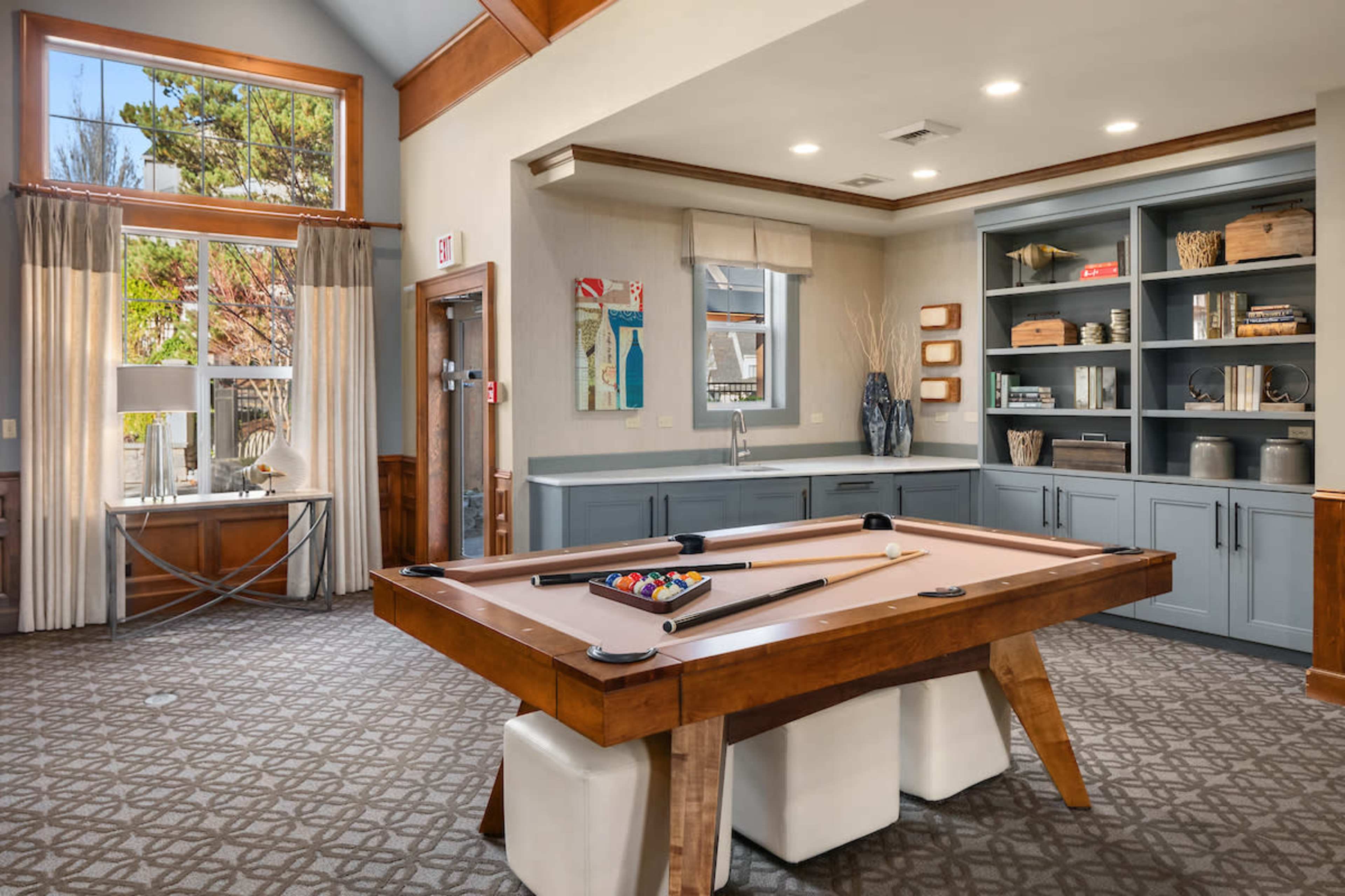 The image shows a room featuring a billiards table at the center, surrounded by shelves with decorative items and large windows allowing natural light to enter.