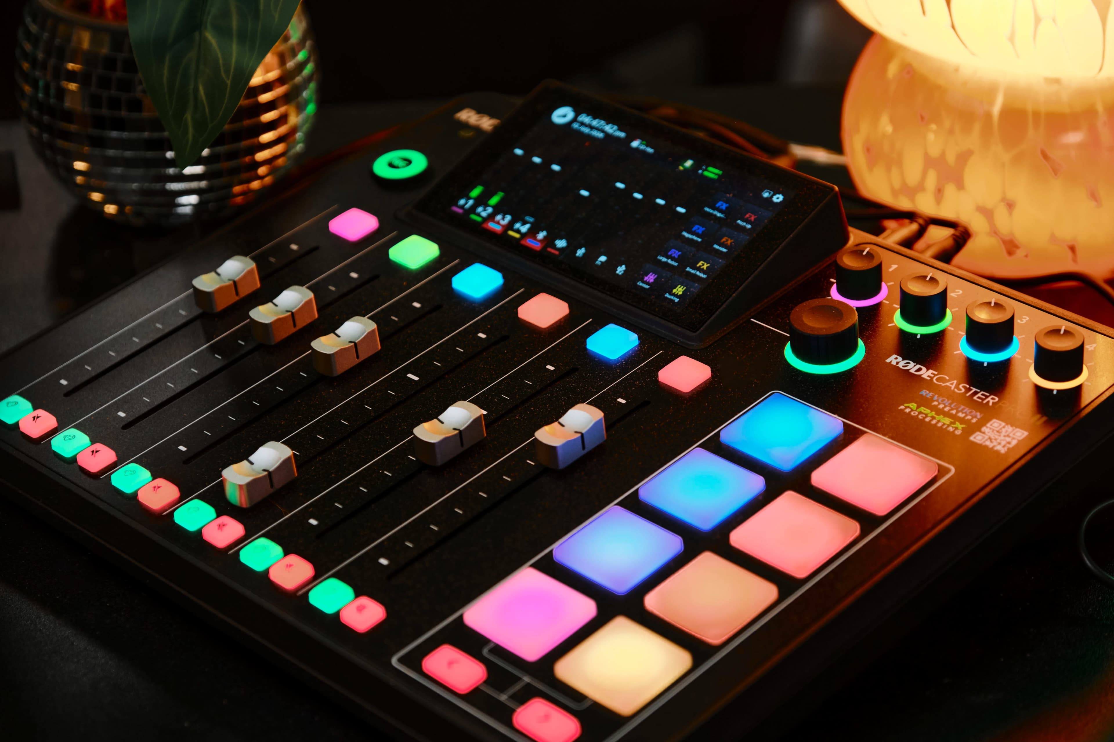 A Rodecaster Pro audio mixing console with colorful buttons and sliders, placed on a dark surface beside a plant and a lamp.