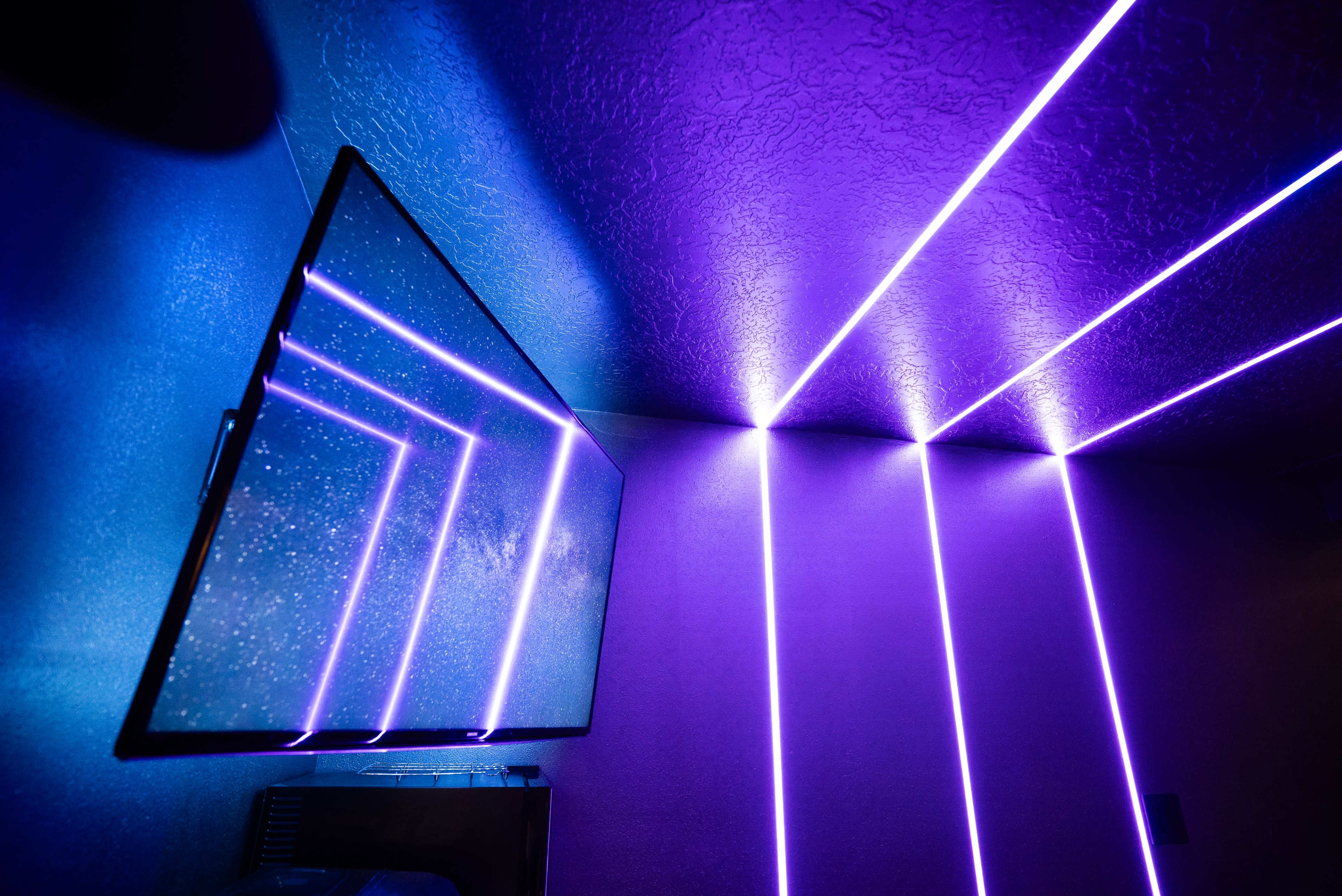 A flat-screen TV is mounted on a wall with purple LED lights creating geometric patterns along the ceiling and wall.