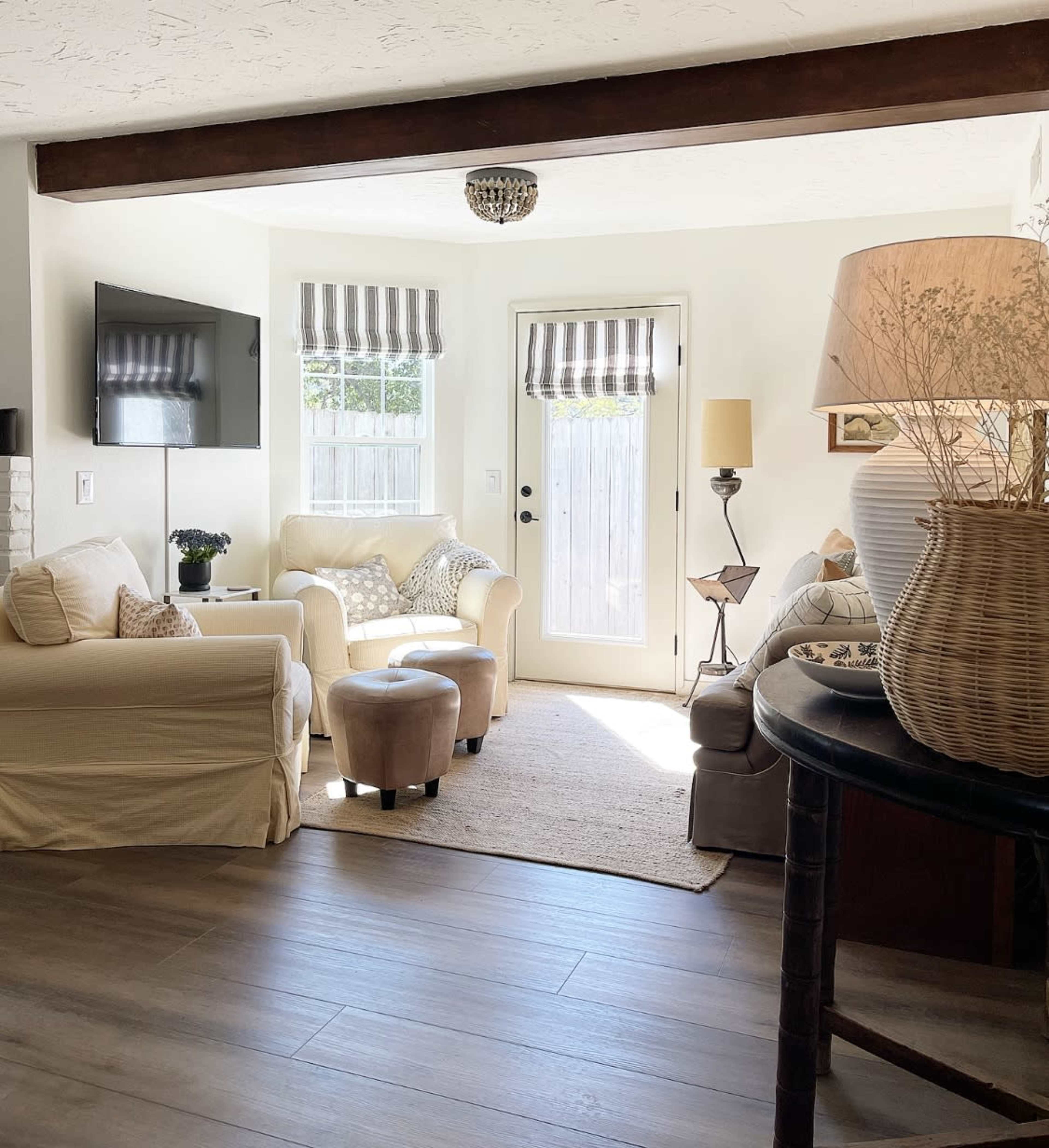 A bright living room features light-colored furniture, a television on the wall, and a door leading to an outdoor area, with decorative striped window treatments and a woven lamp.