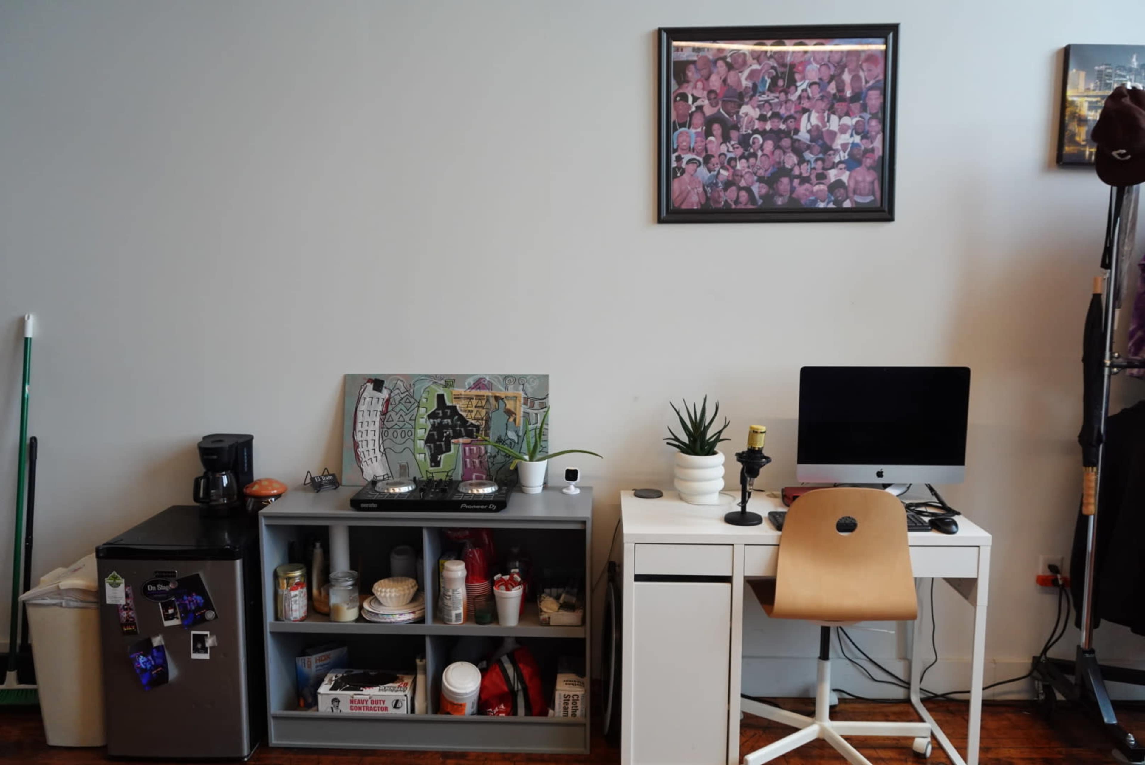 The image shows a small living space with a refrigerator, a shelf containing kitchen items, and a desk with a computer and a chair, all arranged against a light-colored wall.