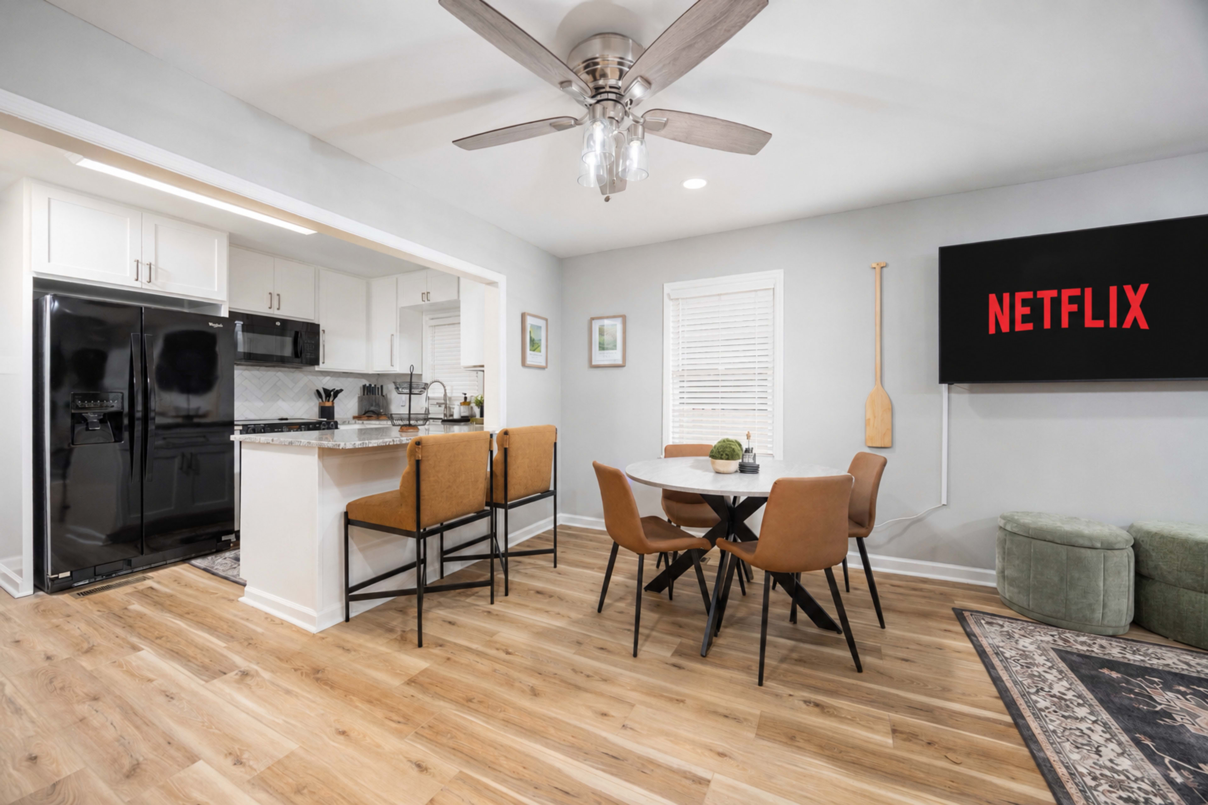 The image shows a modern kitchen and dining area featuring a round table with chairs, a black refrigerator, and a television screen displaying the Netflix logo.