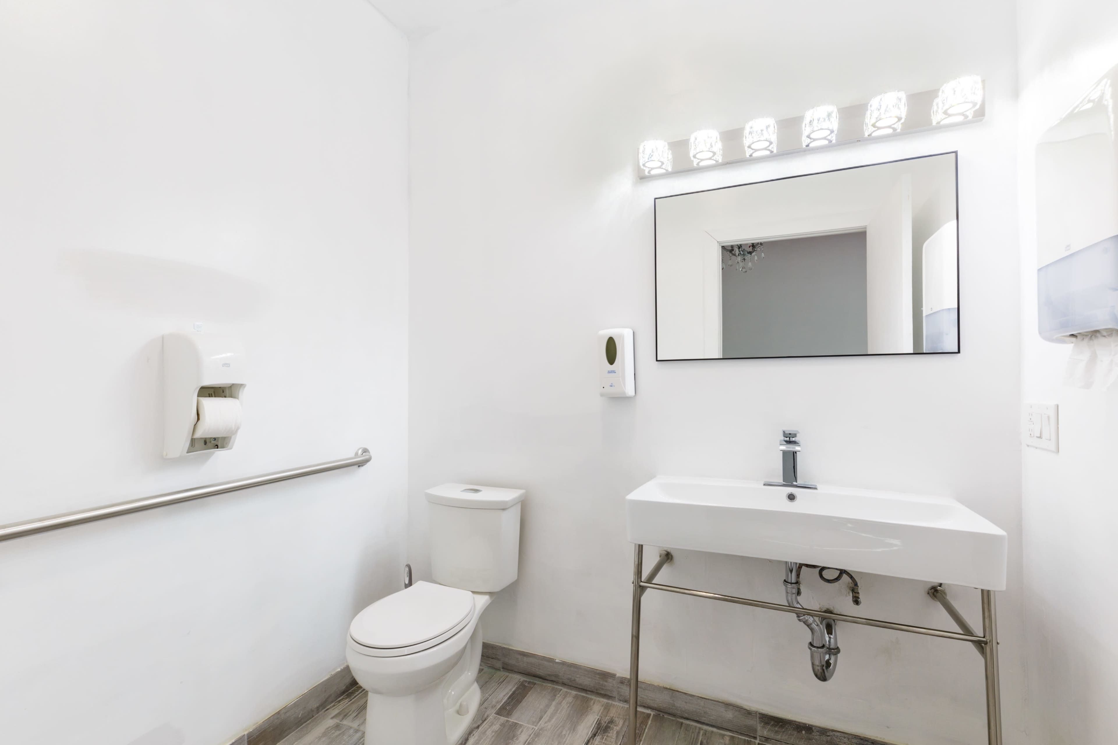 A clean, modern bathroom featuring a toilet, a sink with an undermount design, a large mirror, and basic hygiene fixtures against white walls.