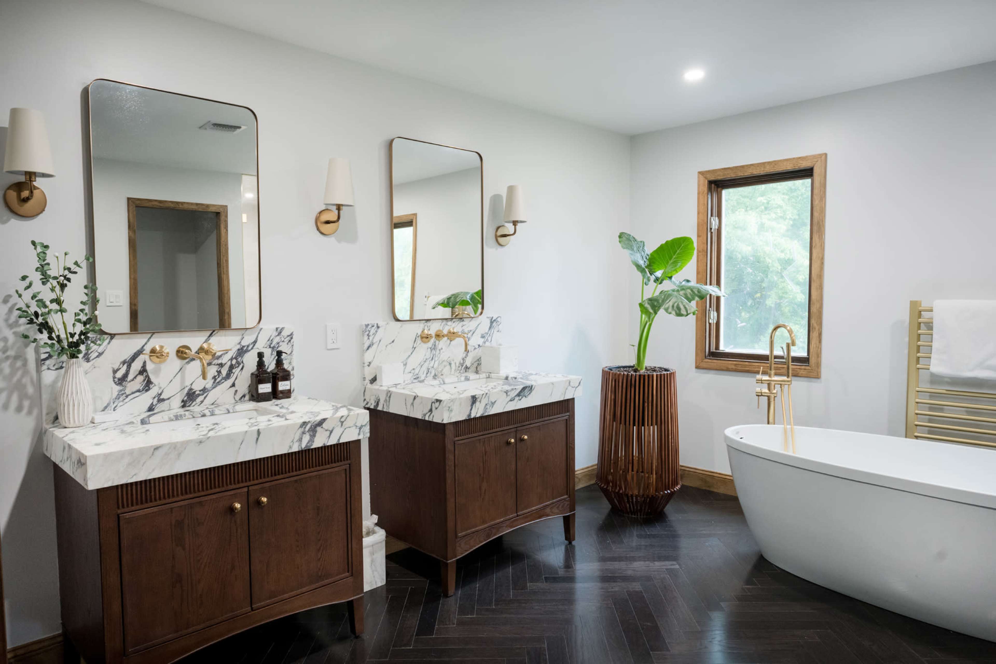 The image depicts a modern bathroom featuring two wooden vanities with marble countertops, a freestanding bathtub, and large windows allowing natural light.