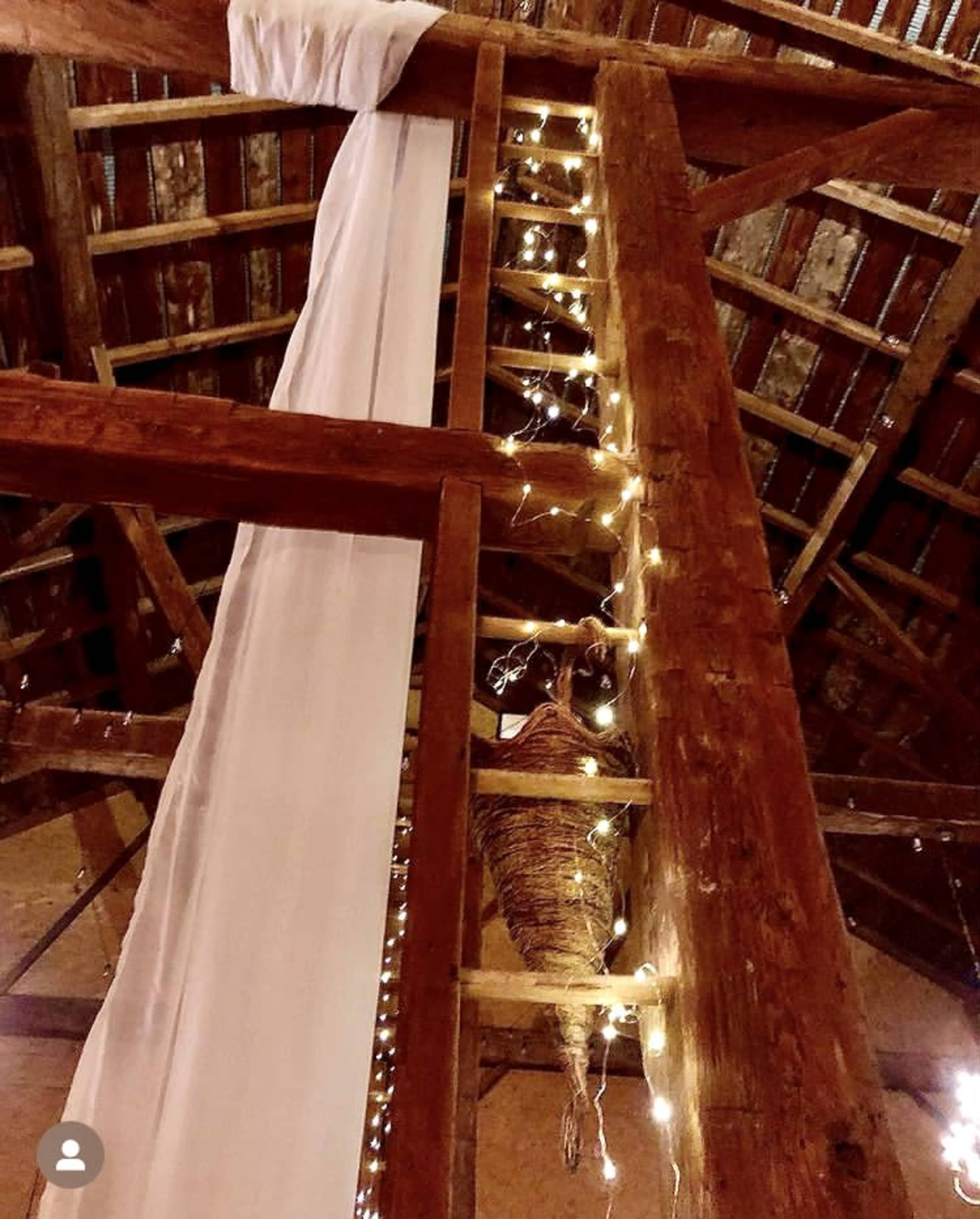 A wooden ladder is positioned against a rustic beam adorned with soft drapery and string lights in a barn setting.