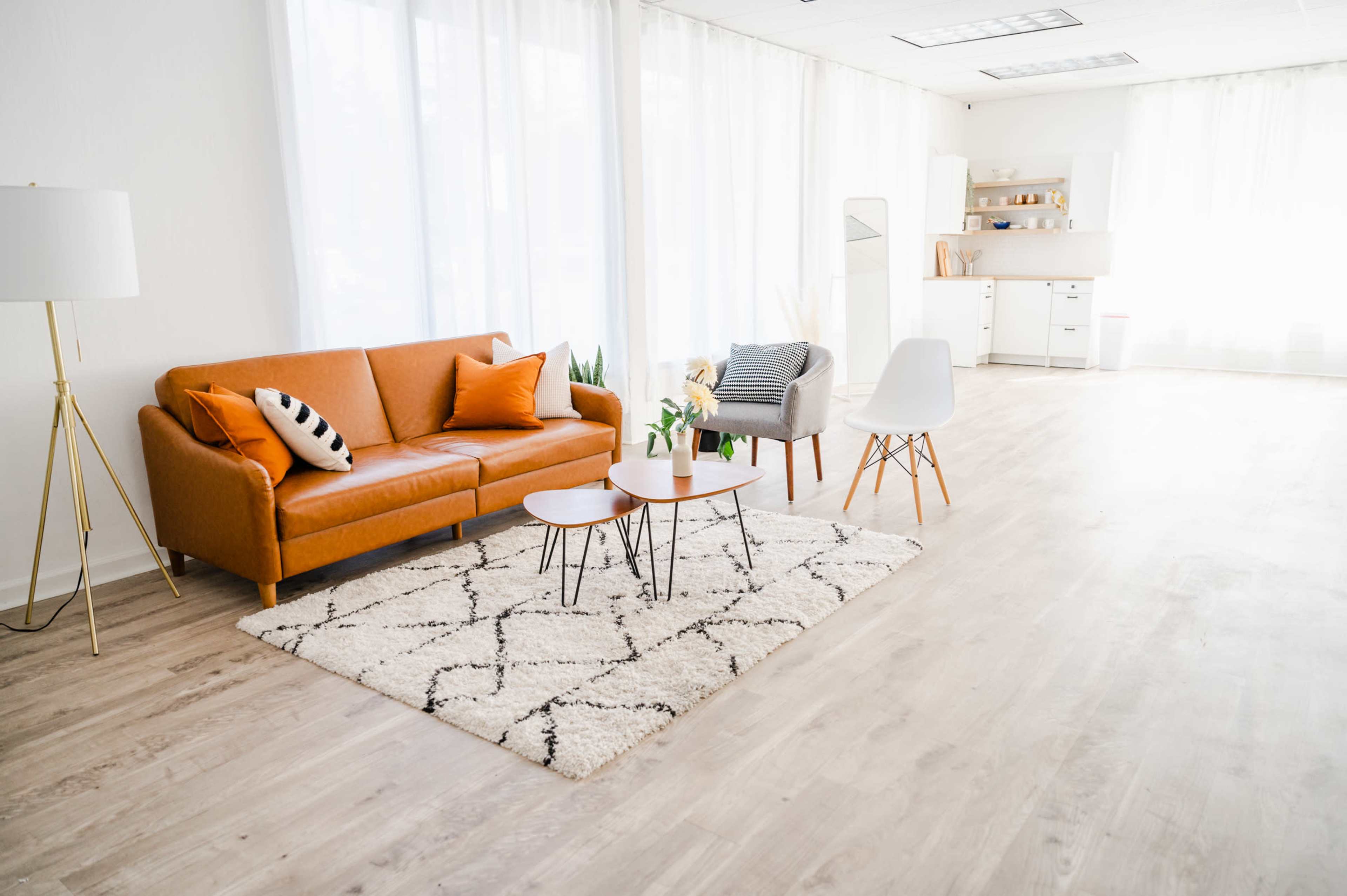 A bright, modern living space featuring an orange sofa, two coffee tables, and a mix of decorative chairs, with large windows allowing natural light to fill the room.