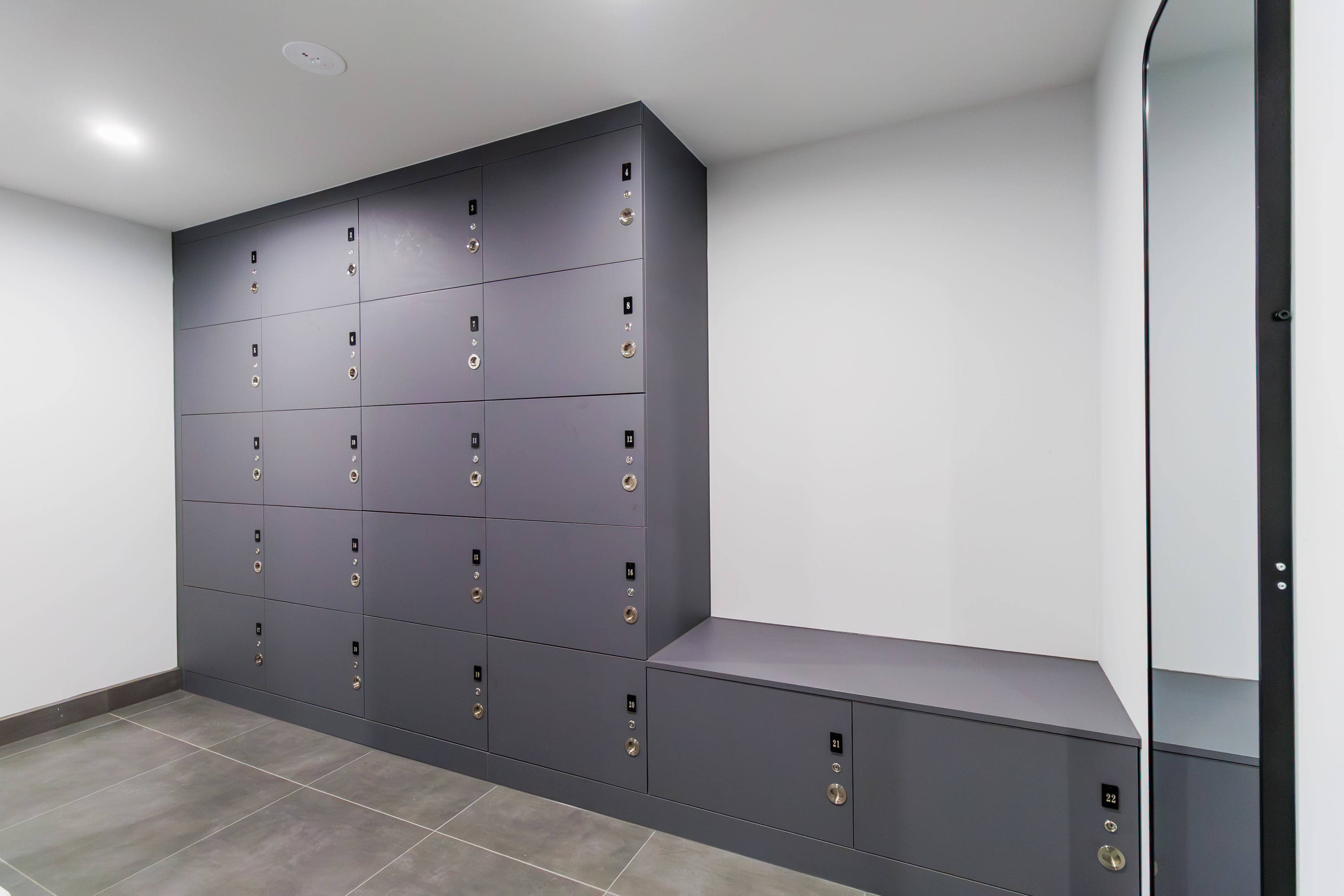 The image shows a row of gray lockers mounted against a white wall, with a bench and a mirror nearby.