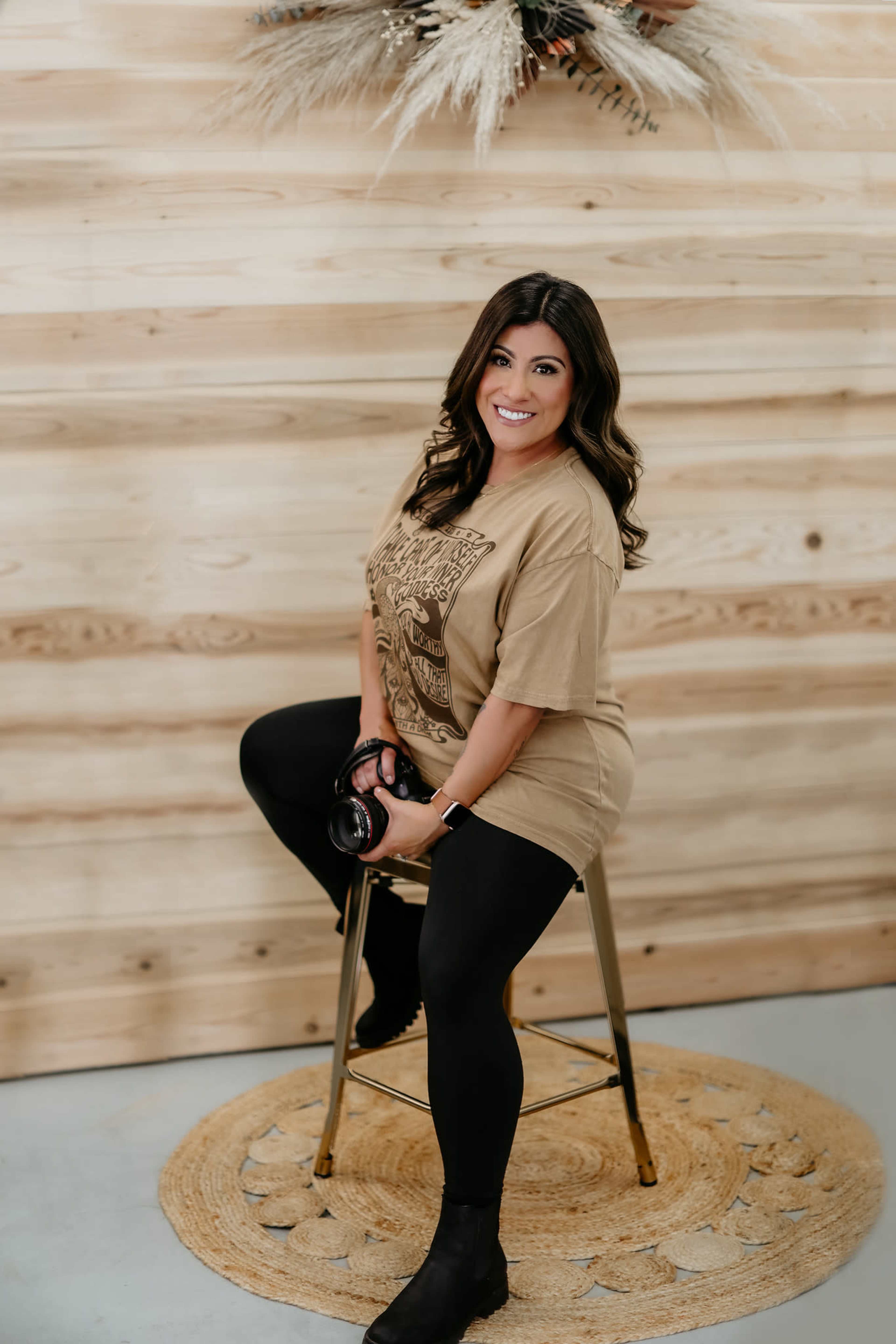 A woman sits on a golden stool, wearing a beige shirt and black leggings, with a pair of headphones around her neck, against a backdrop of wooden paneling.