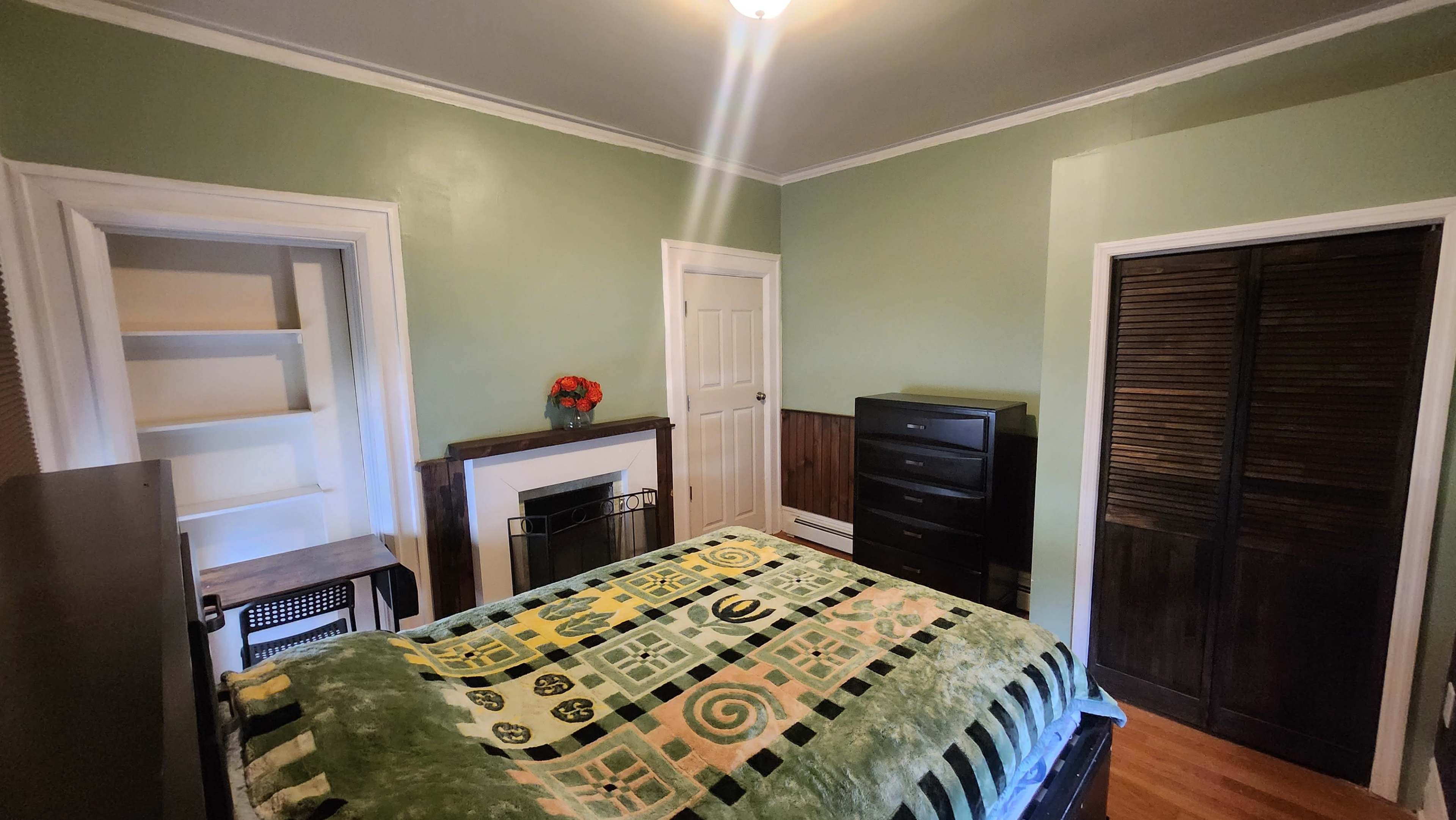 A small bedroom with a bed covered by a green and patterned blanket, two closets, and a dresser against the opposite wall.