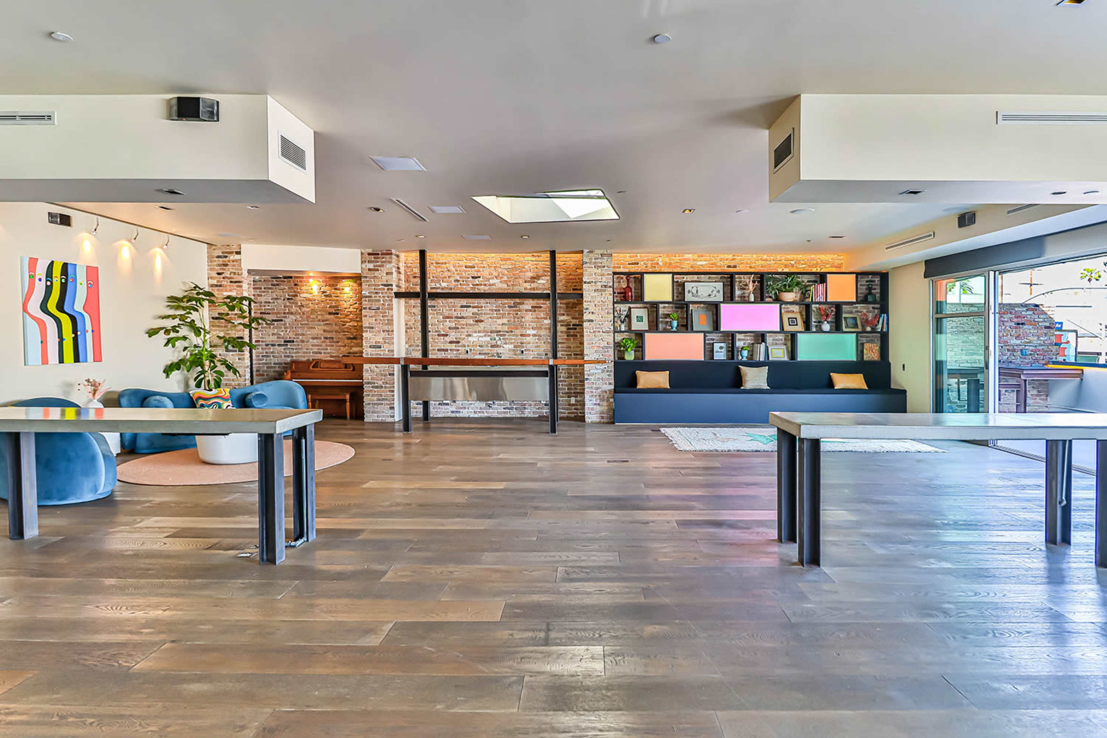 A spacious interior featuring wooden floors, blue seating, a brick wall, and large windows allowing natural light.