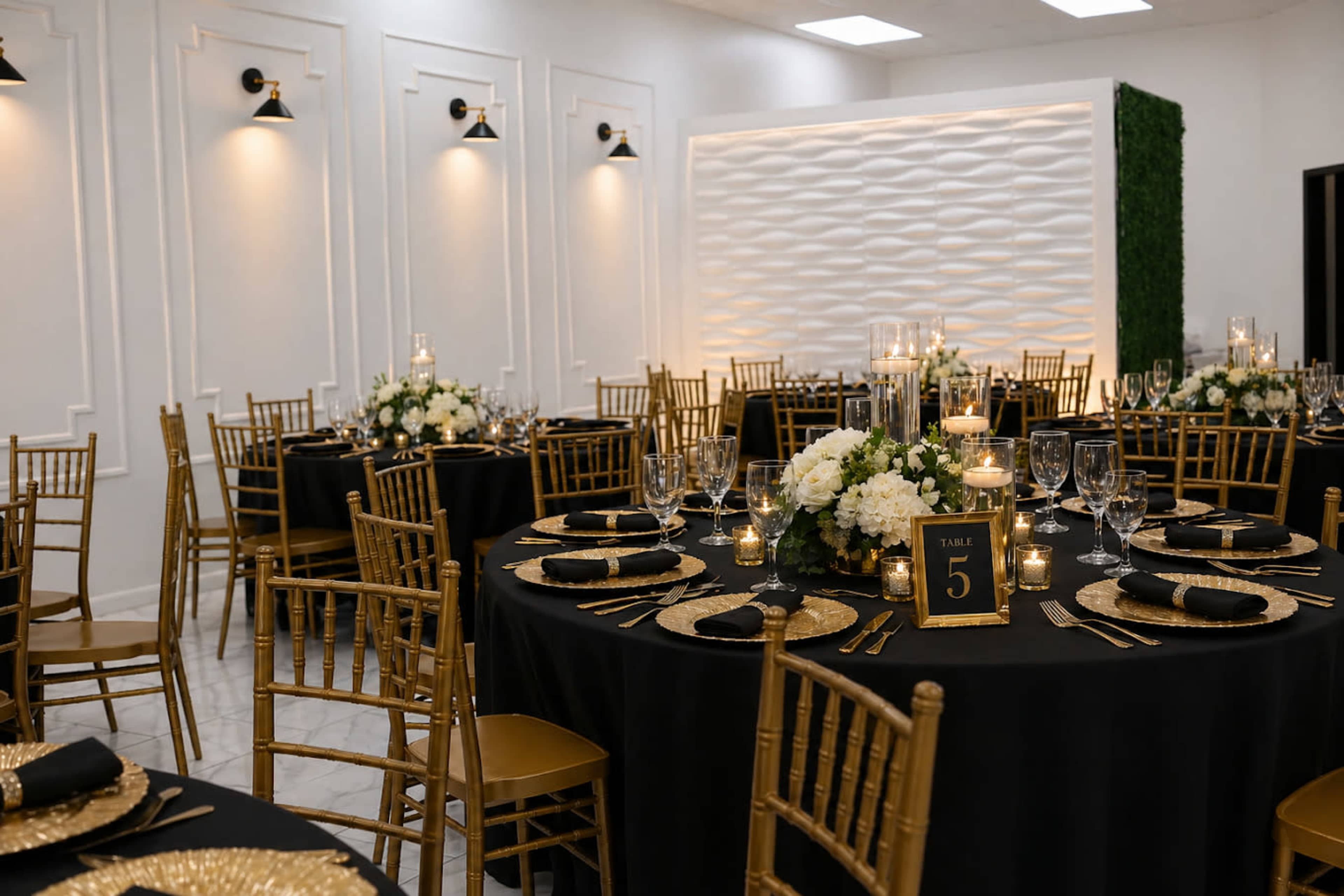 The image shows an elegantly decorated event space with round black tables adorned with gold accents, centerpieces of white flowers, and candlelight, set against a backdrop of textured white walls and greenery.